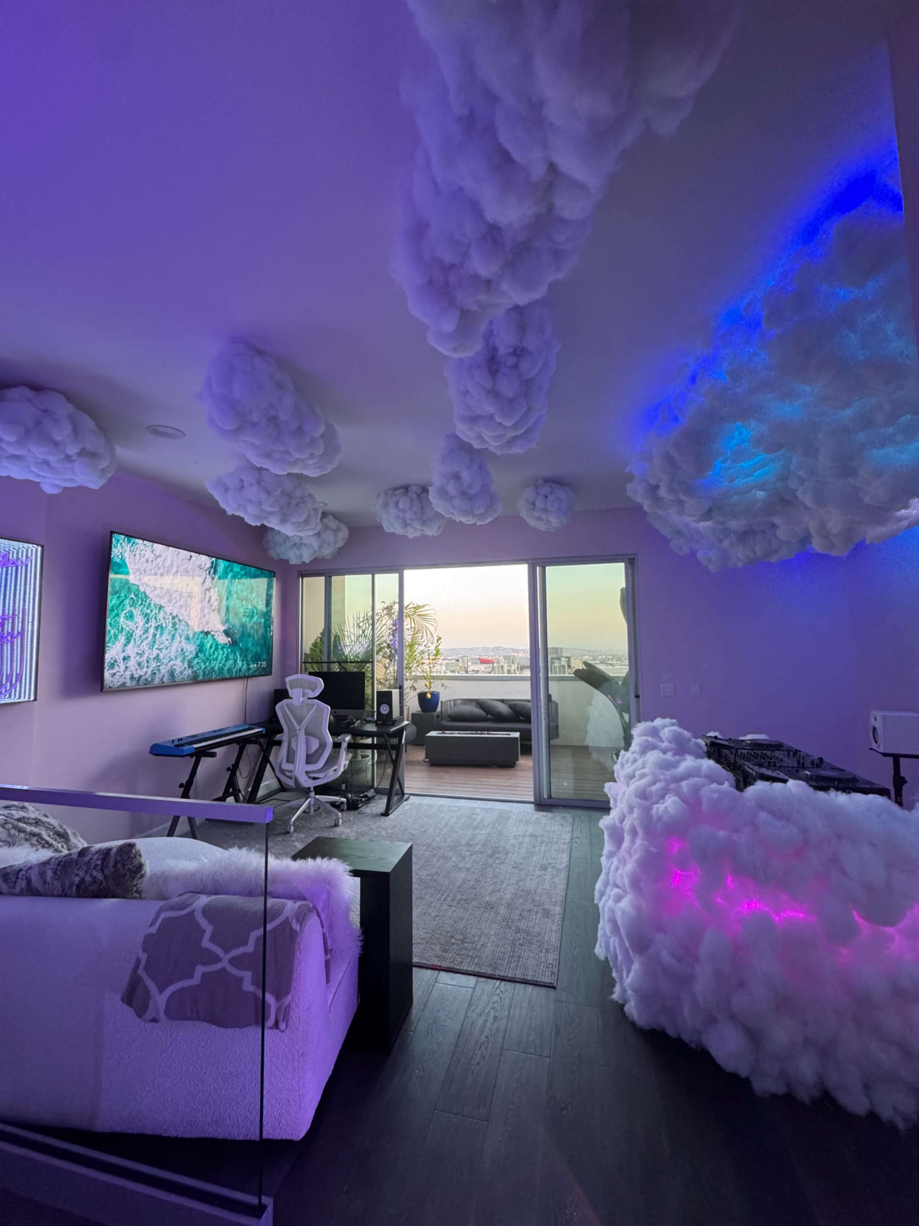 A modern bedroom with a bed, a sectional sofa, and a large television, featuring cloud-like decorations on the ceiling and a scenic view of the outdoors through glass doors.