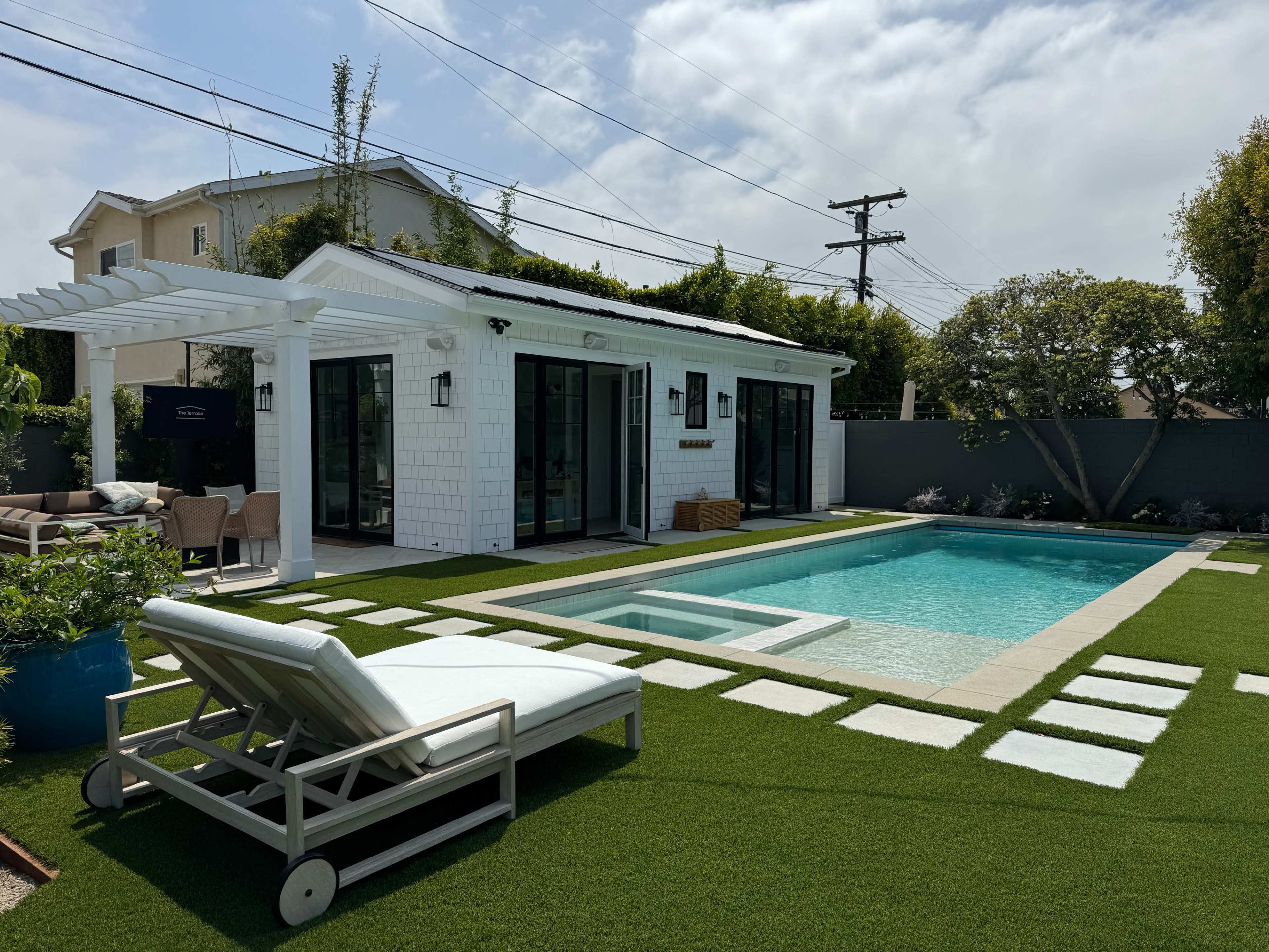 The image shows a modern backyard featuring a swimming pool, a lounge chair, a covered patio, and well-manicured grass with stone pathways.