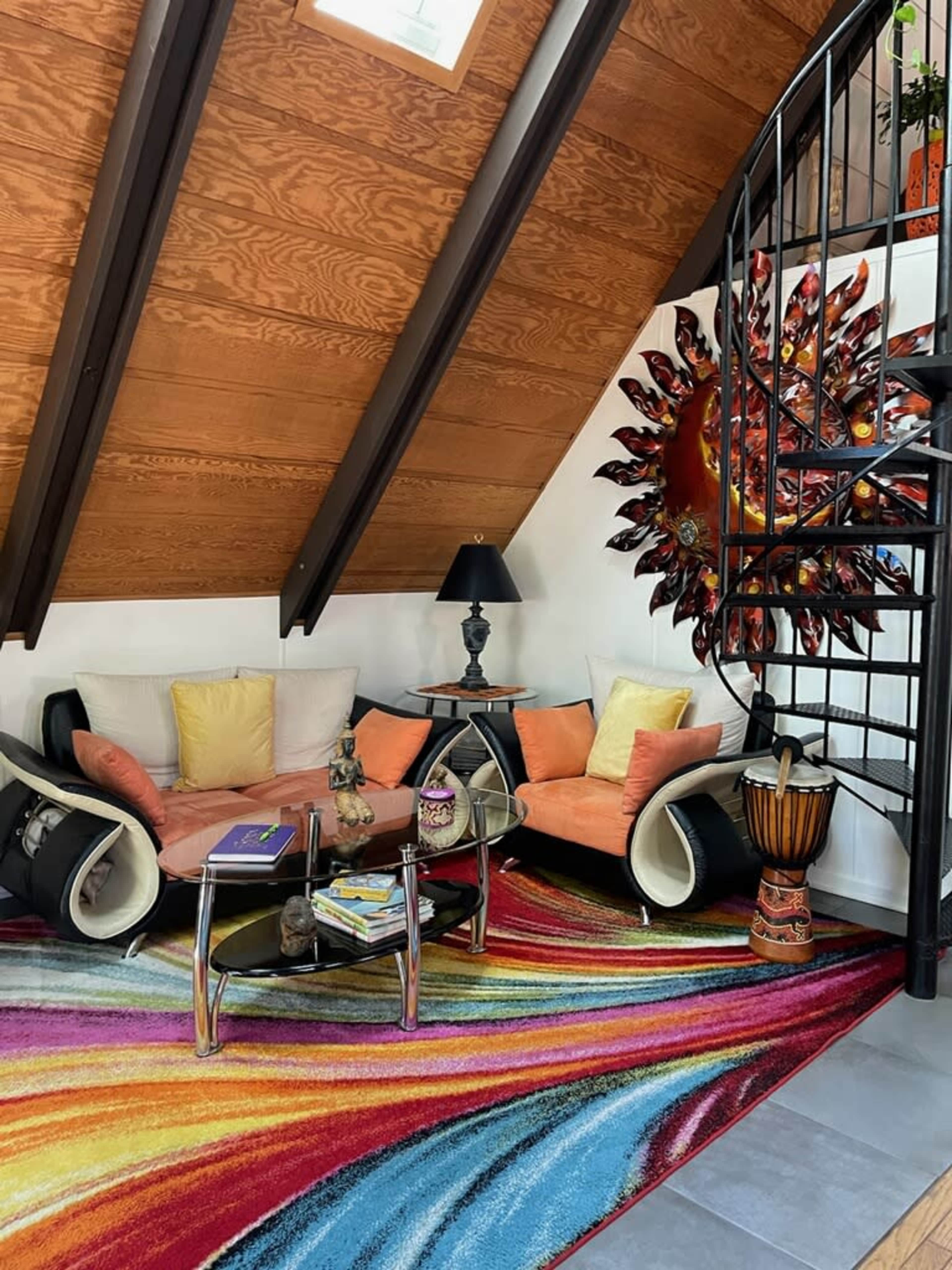 A cozy living room with a colorful area rug, a circular glass coffee table, two orange sofas, and an ornate wall decoration, all under a slanted wooden ceiling.