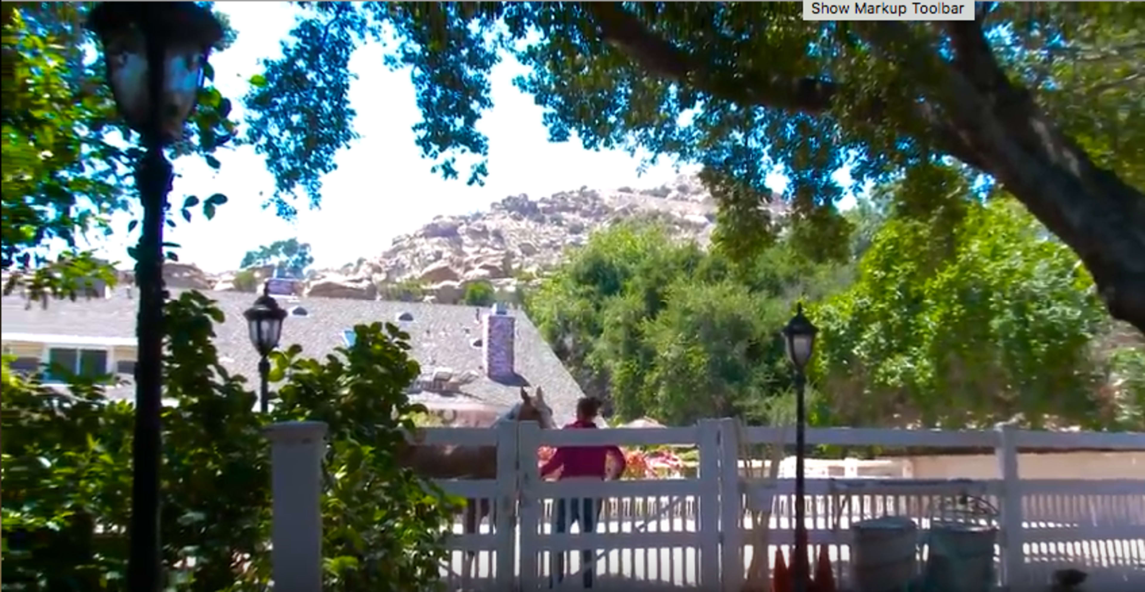 A person stands near a white fence in front of a house, with a rocky hill and trees in the background.