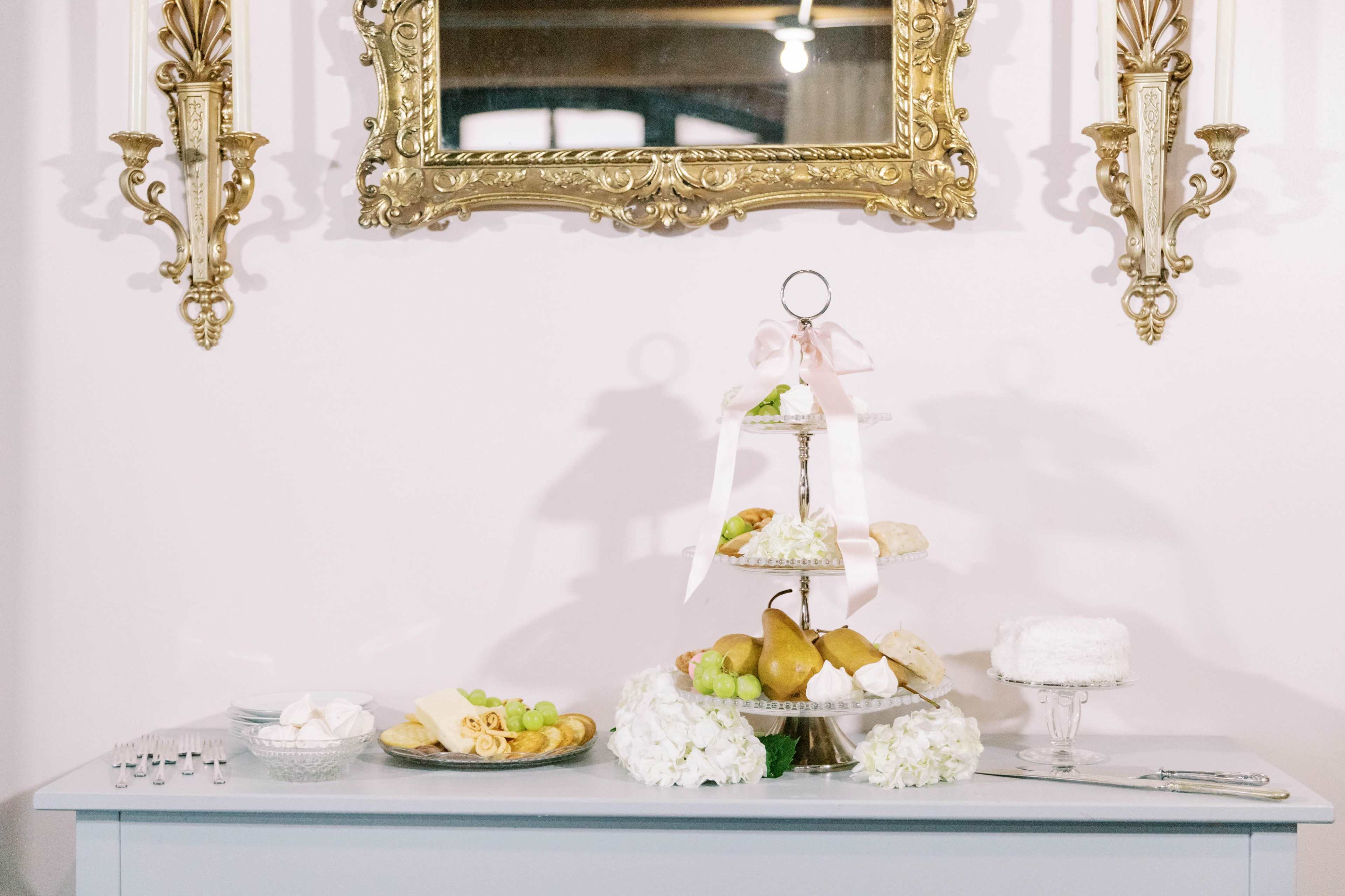 A tiered stand filled with pastries and fruits is displayed on a light-colored table beneath an ornate mirror and golden wall sconces.