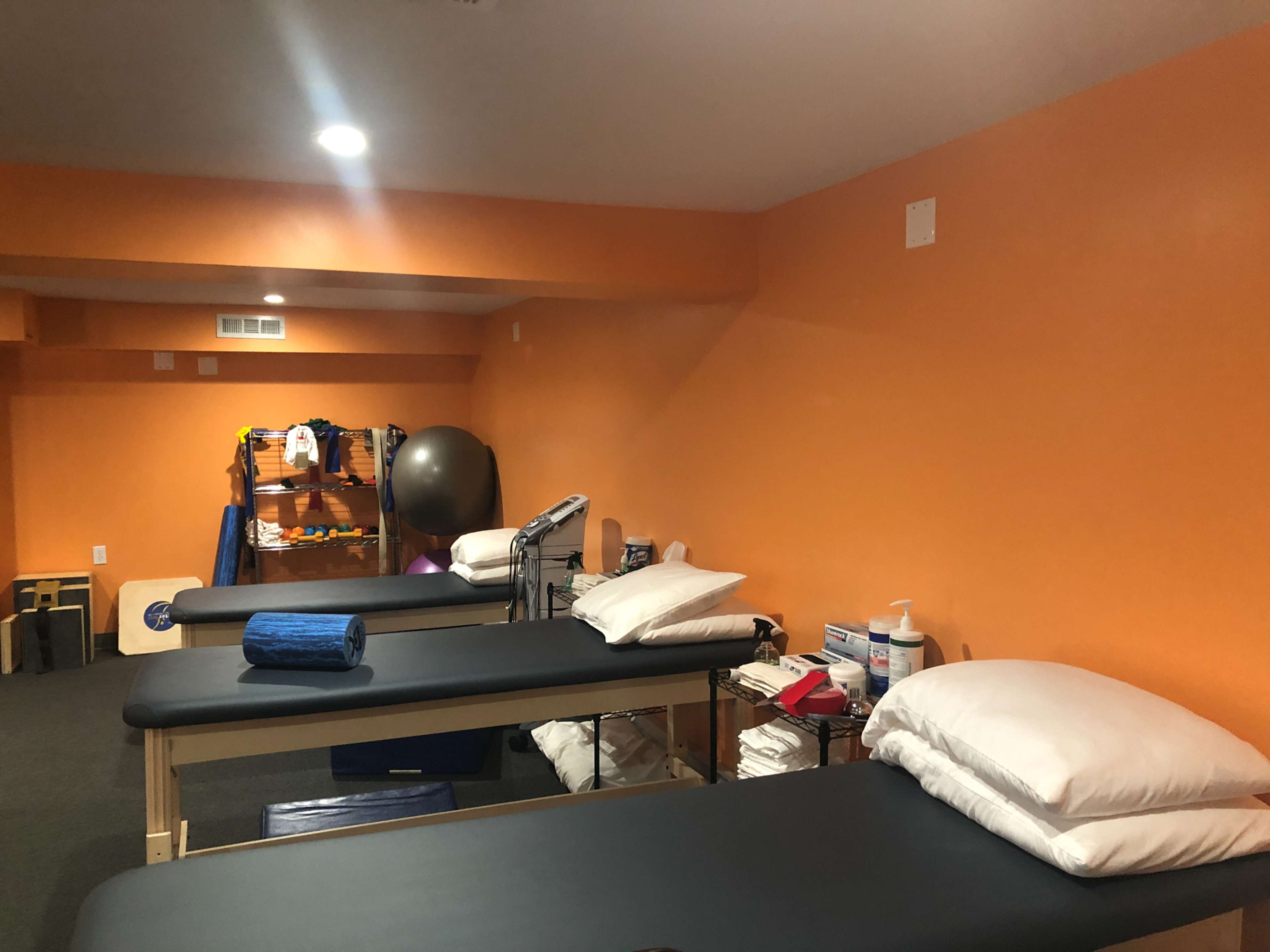 The image shows a room with several massage tables, orange walls, and various exercise equipment in a therapy or fitness setting.