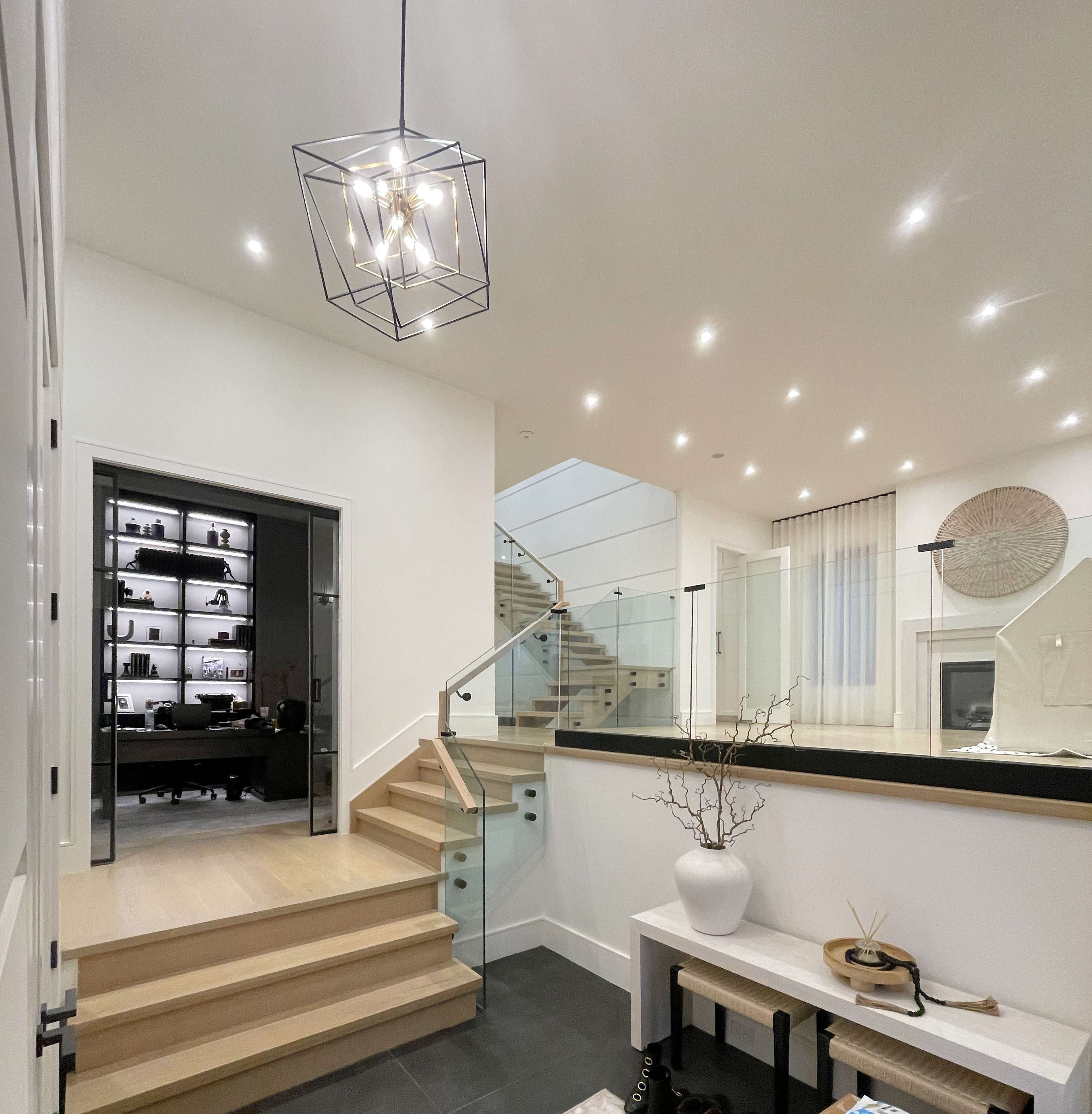 A modern entrance hall with a staircase leading to an upper level, a geometric pendant light, and a workspace visible in the background.