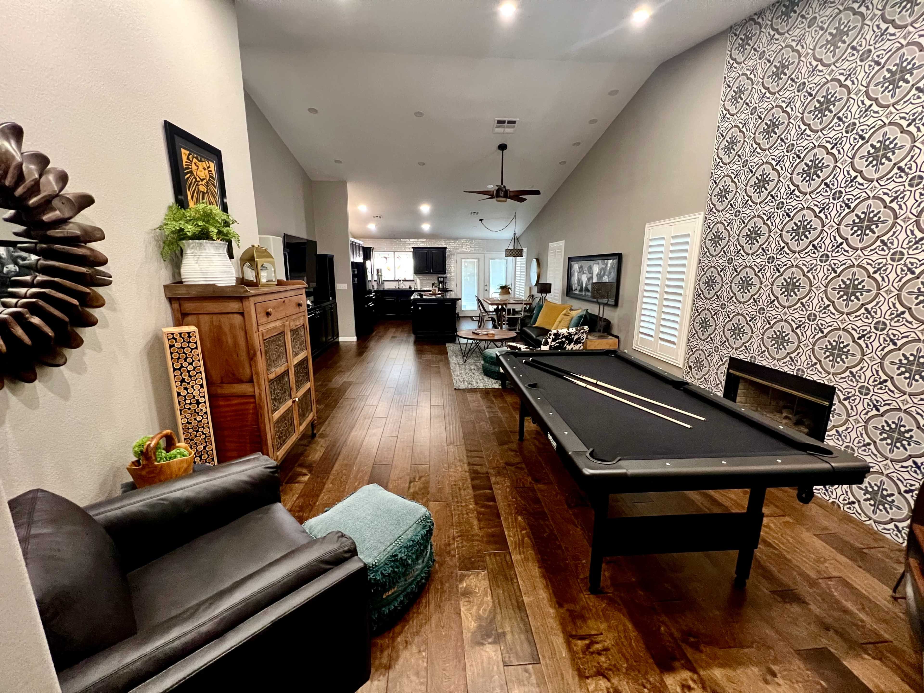 The image shows a spacious living area featuring a billiard table, dark wood floors, and patterned wallpaper, with a view of a kitchen in the background.