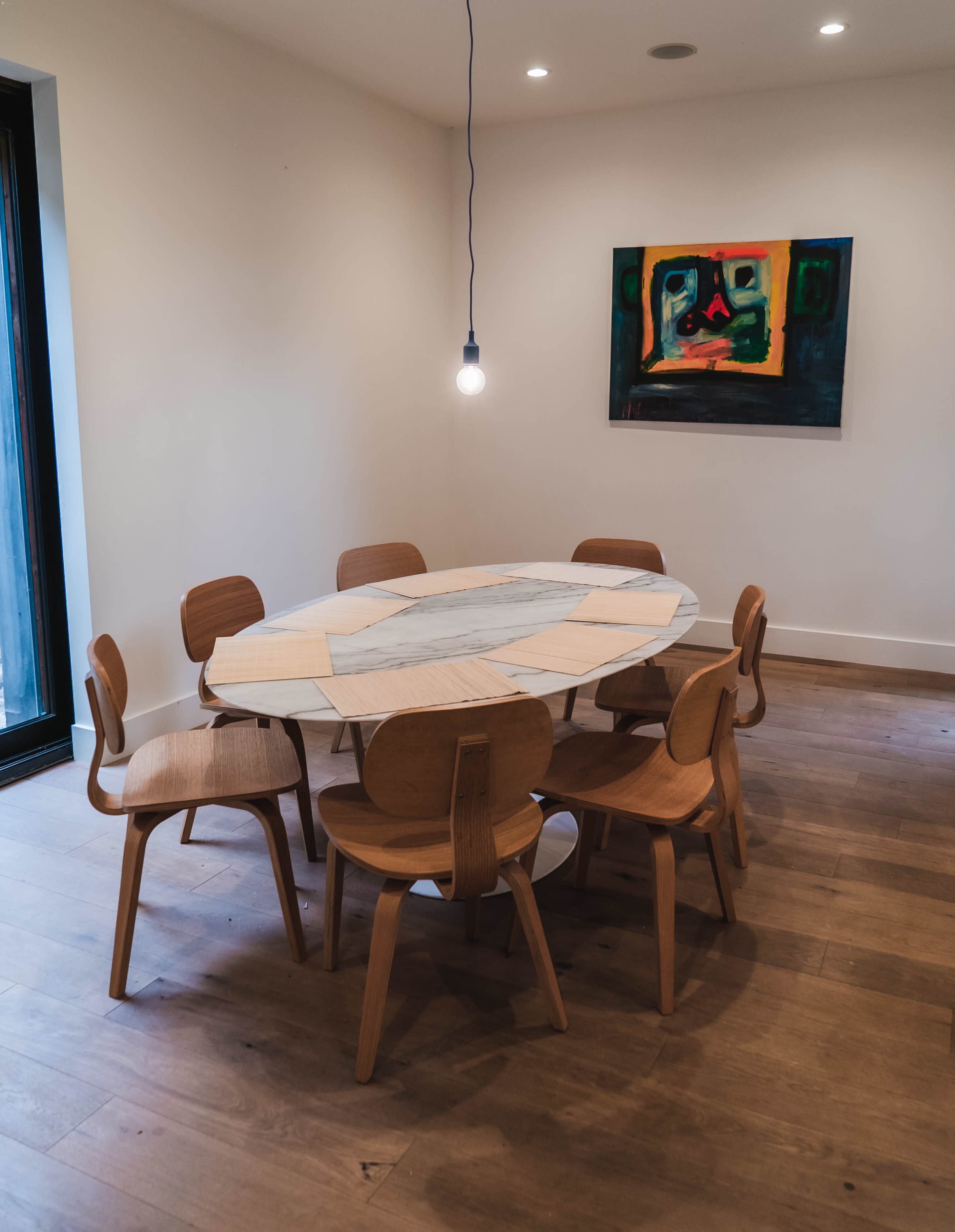 A round marble dining table surrounded by six wooden chairs is set in a minimalist dining area with a wall painting and a single light bulb overhead.