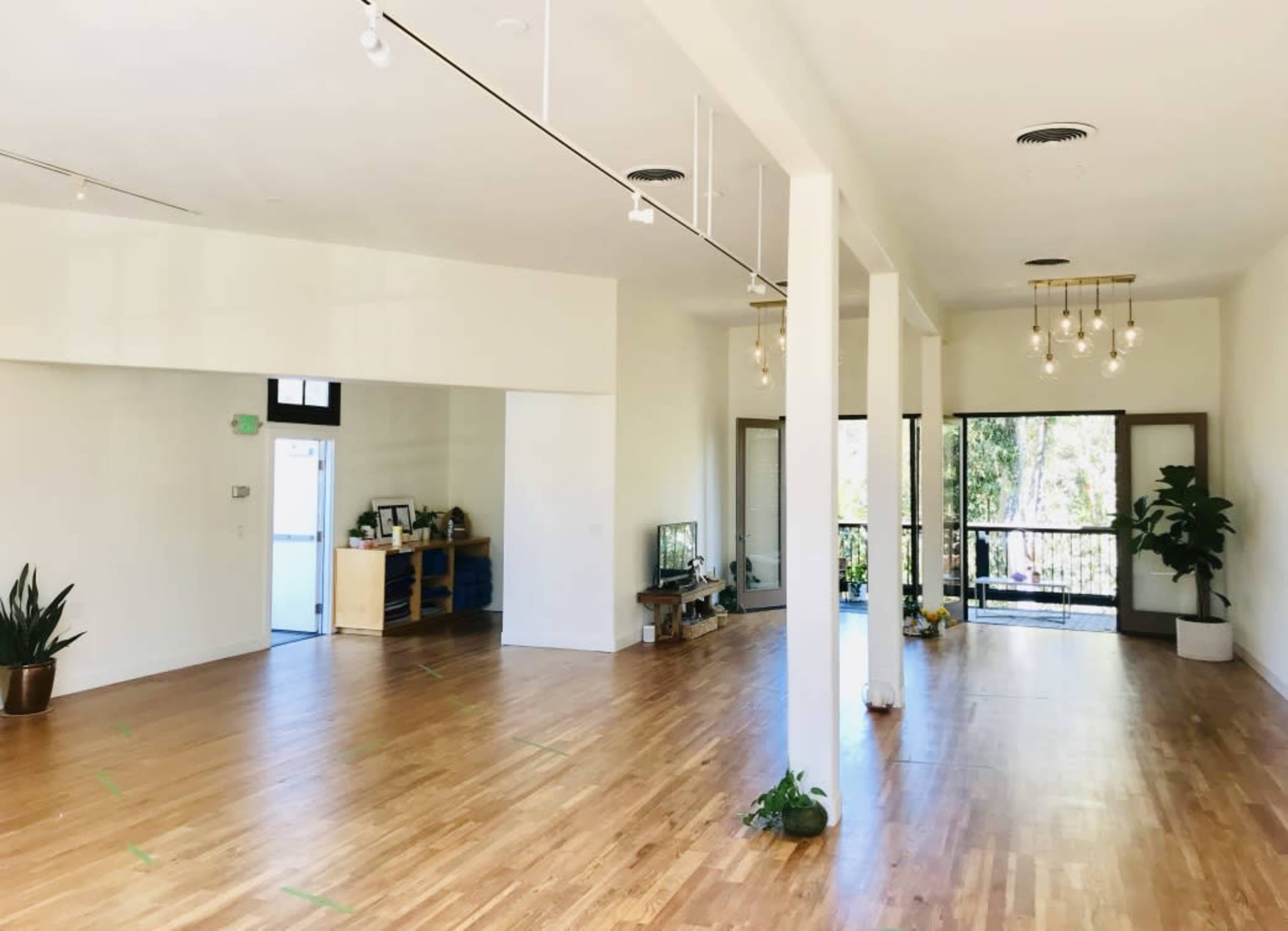 The image shows a spacious, well-lit room with wooden flooring, potted plants, and large windows providing access to an outdoor area.