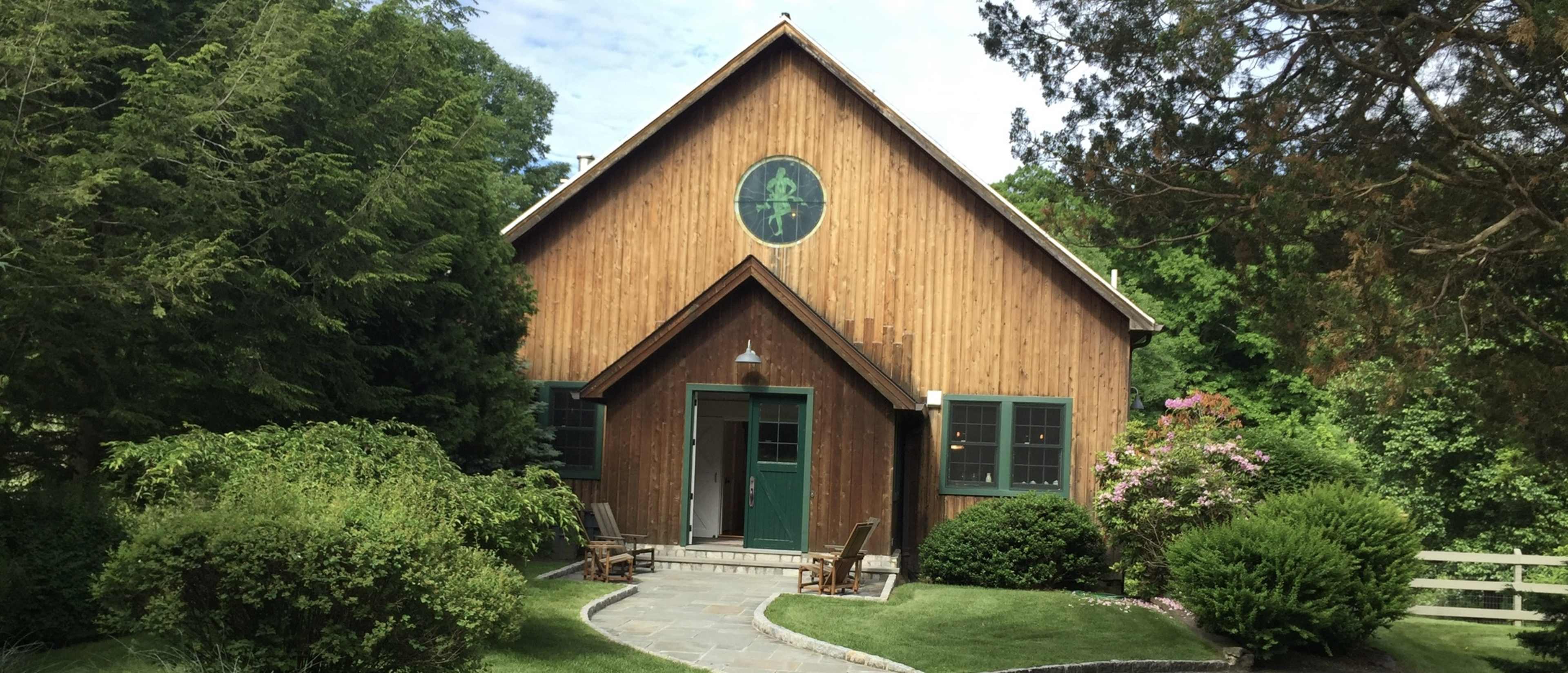 A wooden house with a triangular roof and a circular window stands surrounded by neatly trimmed bushes and a stone pathway.