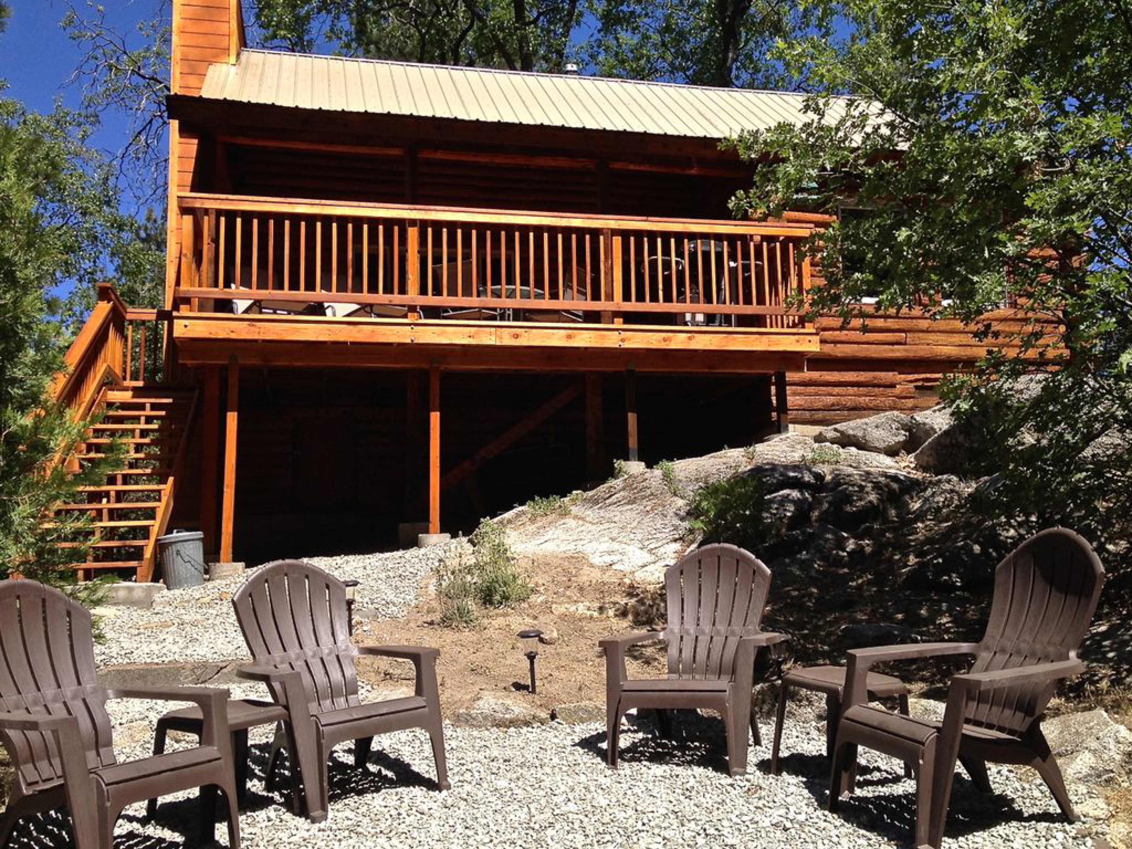 A wooden cabin with a porch is situated on a gravel area, surrounded by rocks and trees, with several chairs arranged in front.