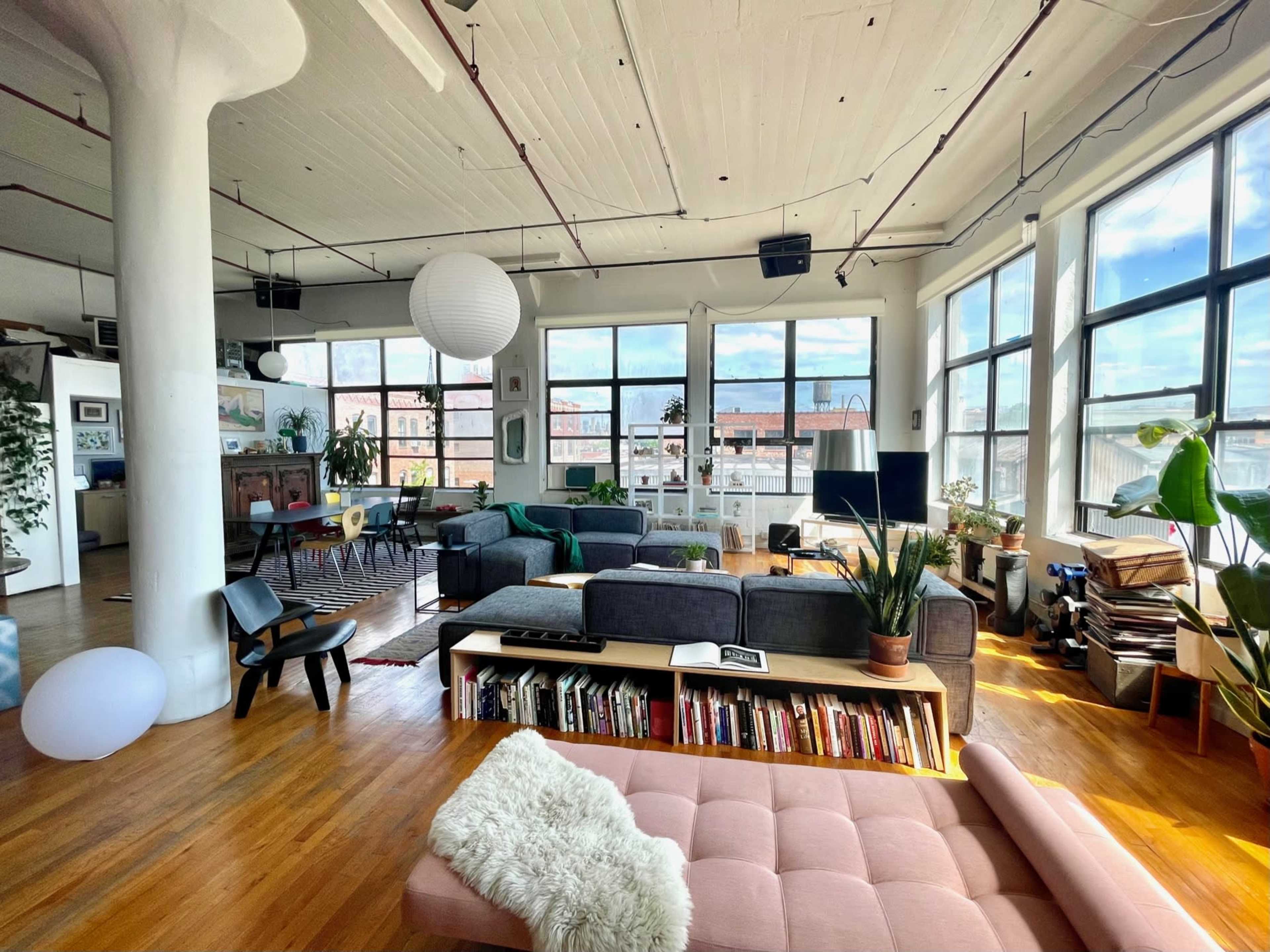 A spacious, sunlight-filled loft features a mix of comfortable seating areas, plants, and large windows with views of the city.
