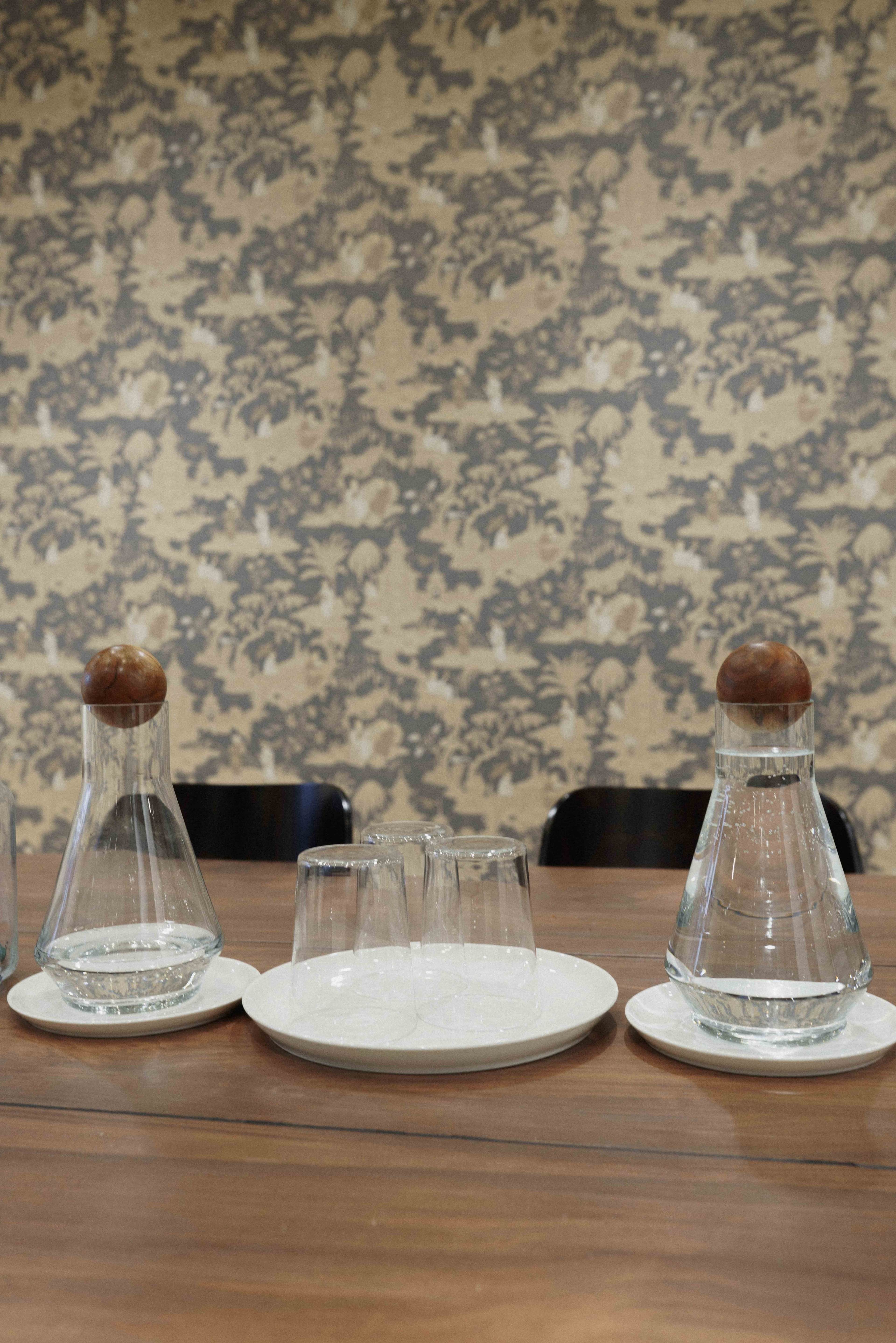 Two glass carafes and three short glasses arranged on a white plate on a wooden table, with a patterned wallpaper in the background.
