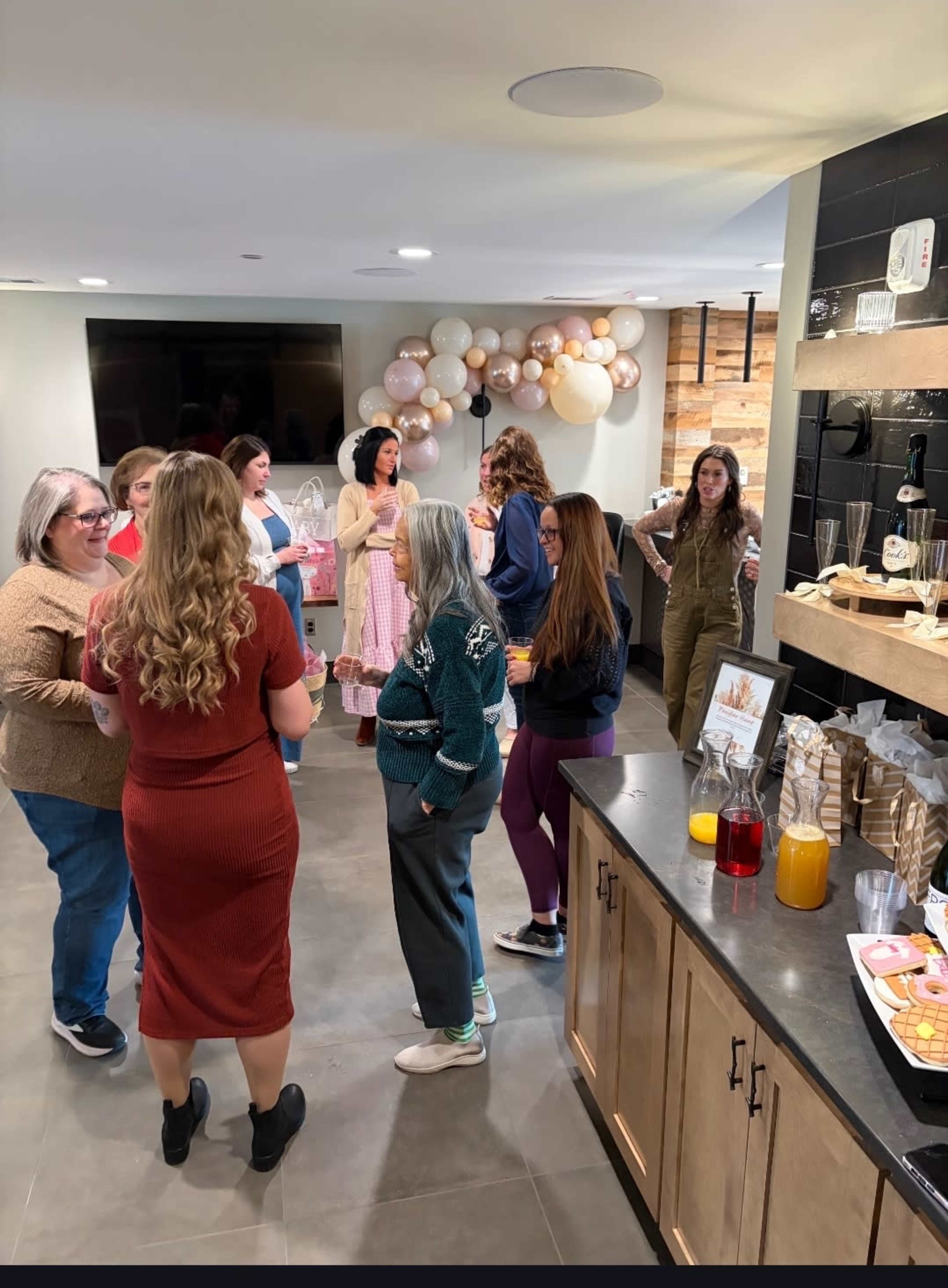 A group of women socialize at a gathering in a modern, decorated indoor space featuring a snack table and decorative balloons.