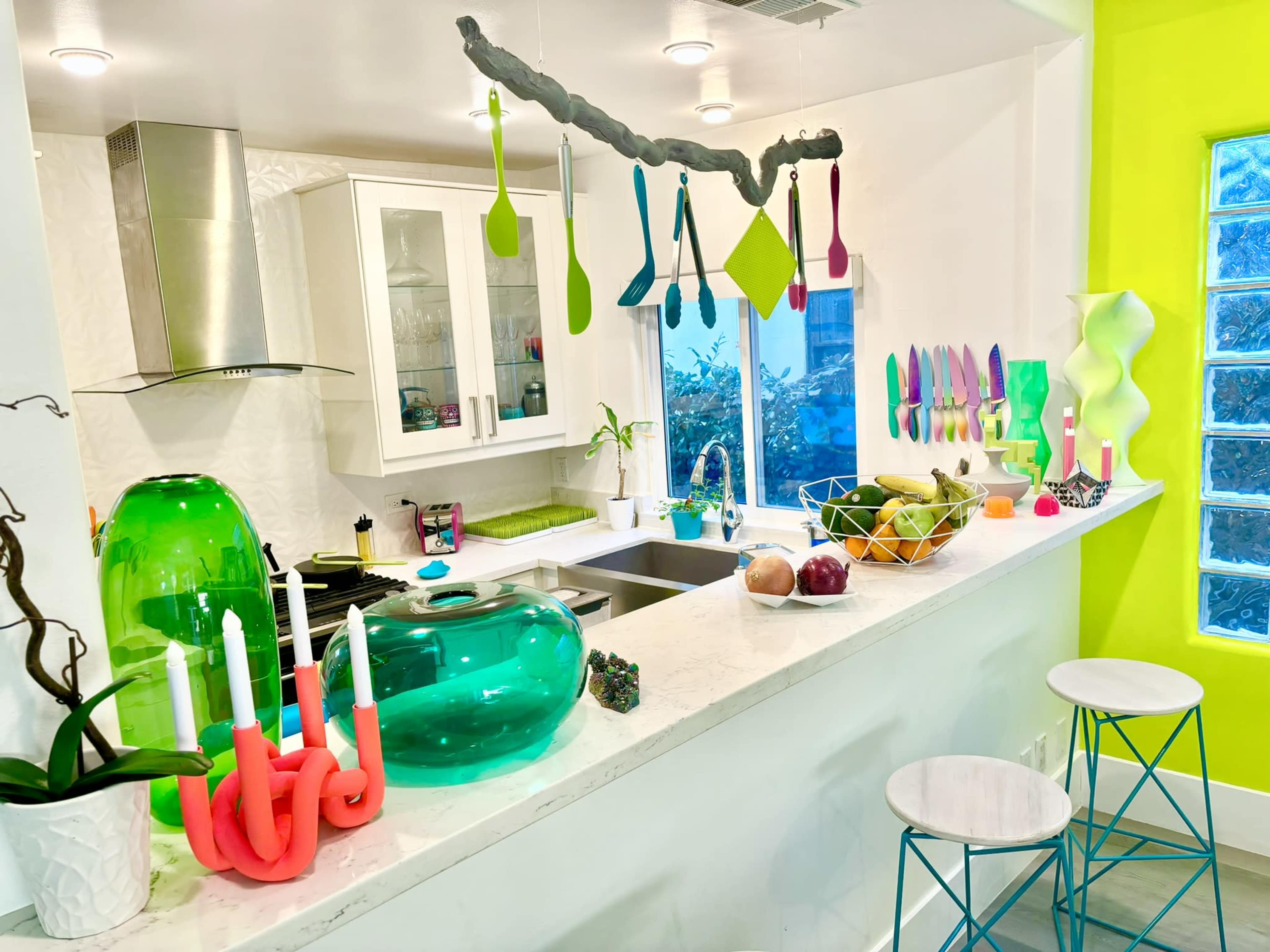 The image shows a modern kitchen with bright green and pink accents, featuring a marble countertop, colorful kitchen utensils hanging above, and various fruits displayed in bowls.