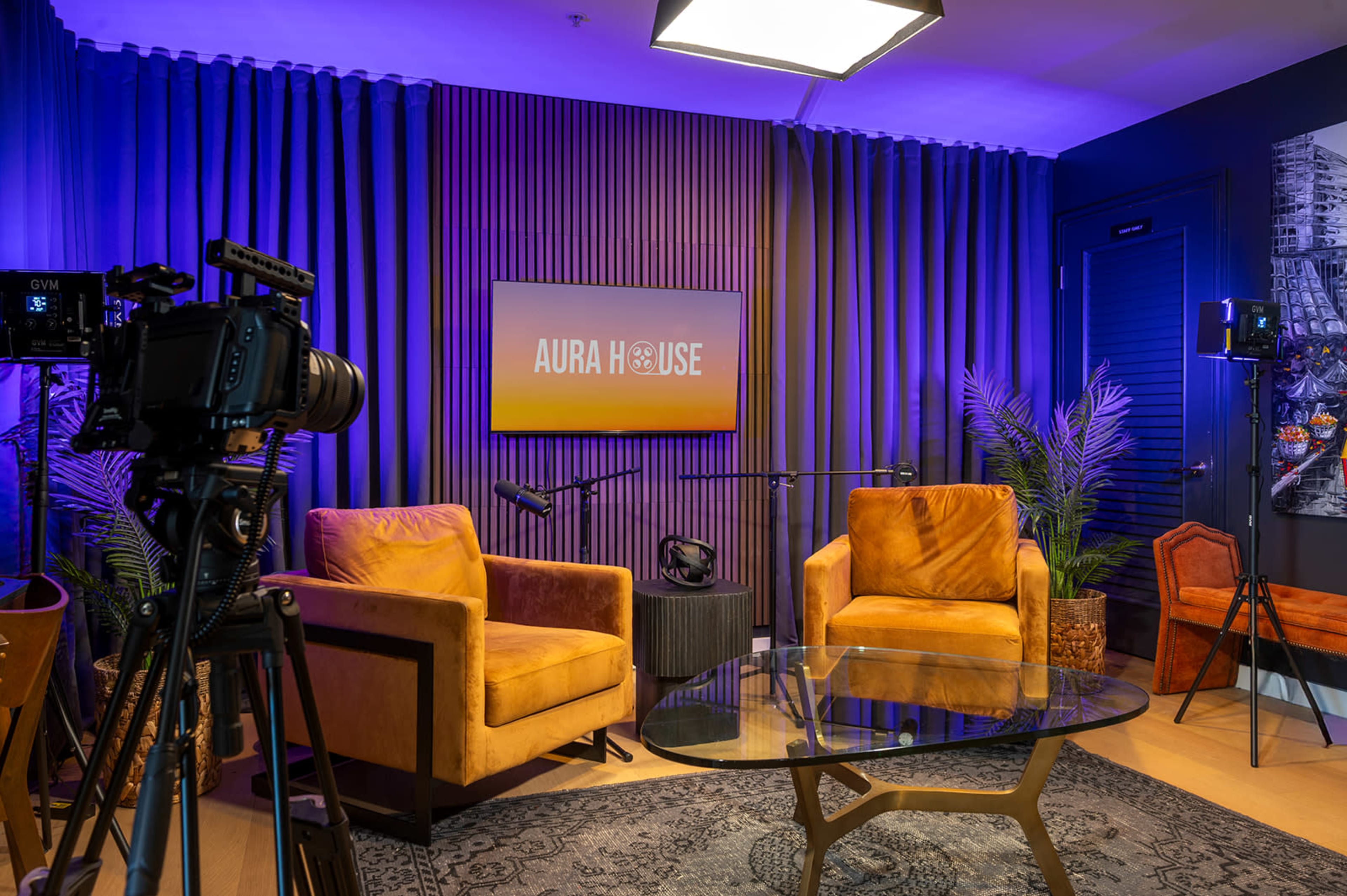 The image shows a modern studio setup with two orange chairs, a glass coffee table, and a large screen displaying "AURA HOUSE" against a backdrop of purple curtains and decorative plants.