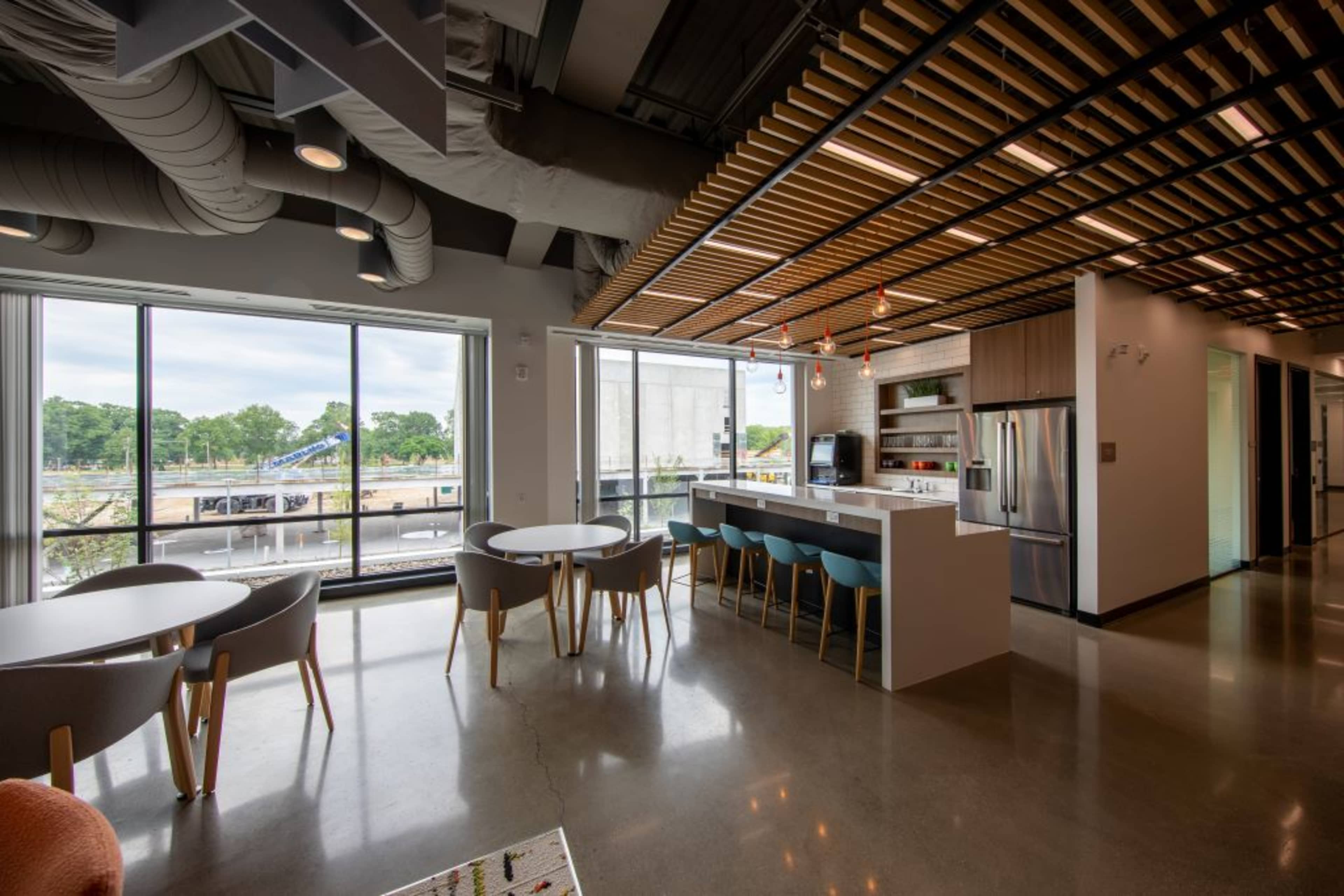 A modern office space featuring a kitchenette area with bar seating, dining tables, and large windows overlooking an outdoor site.