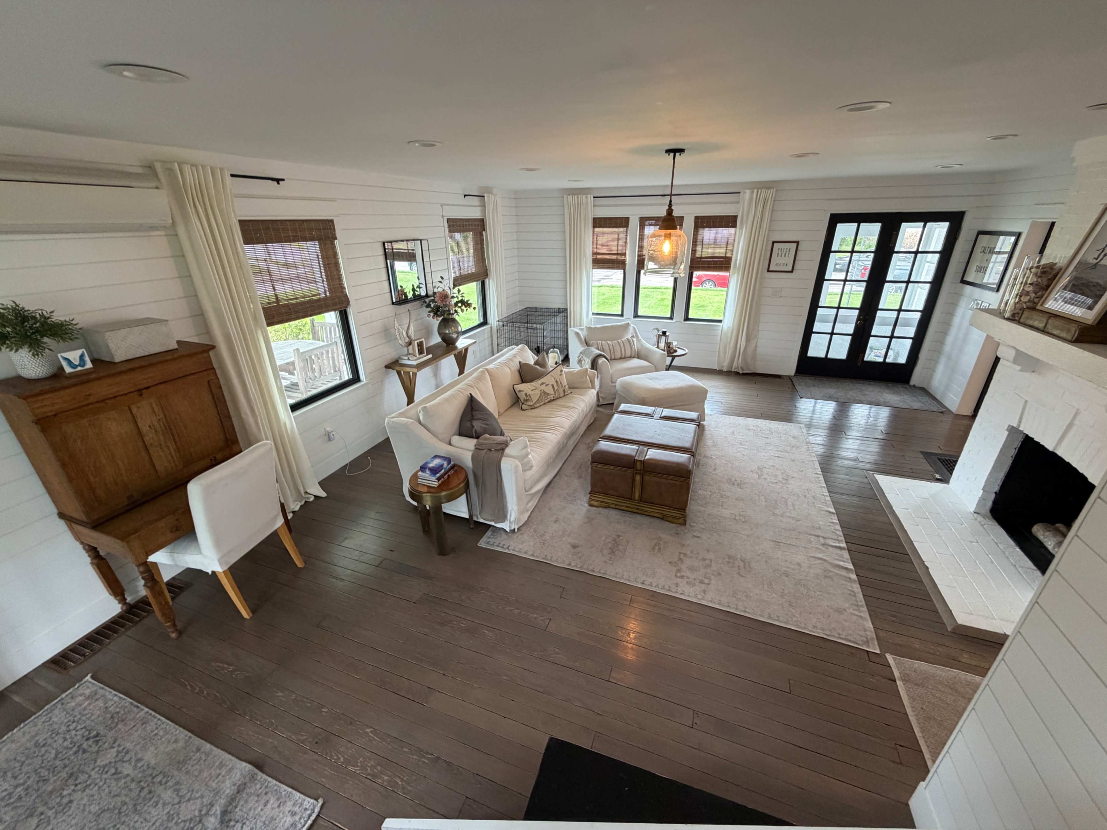 The image shows a bright living room with wooden floors, large windows, a white sofa, a coffee table, and a fireplace.