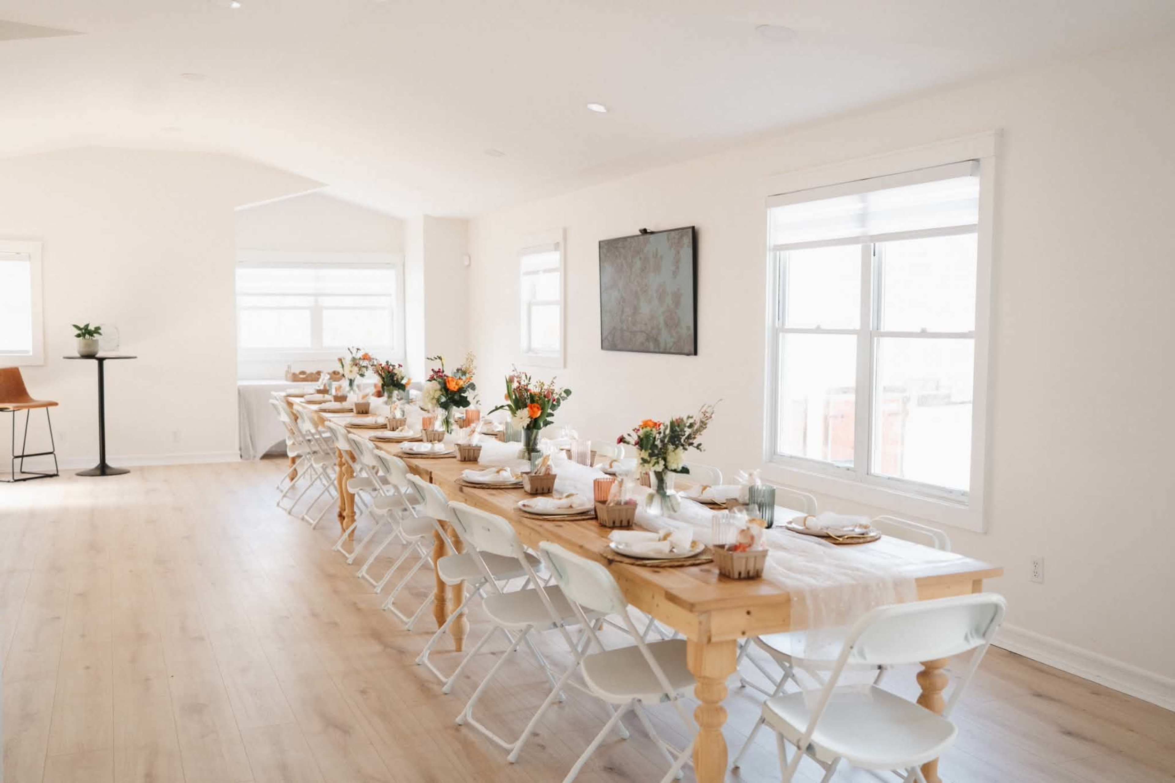 A long, elegantly set dining table with floral centerpieces is arranged in a bright, minimalist room.