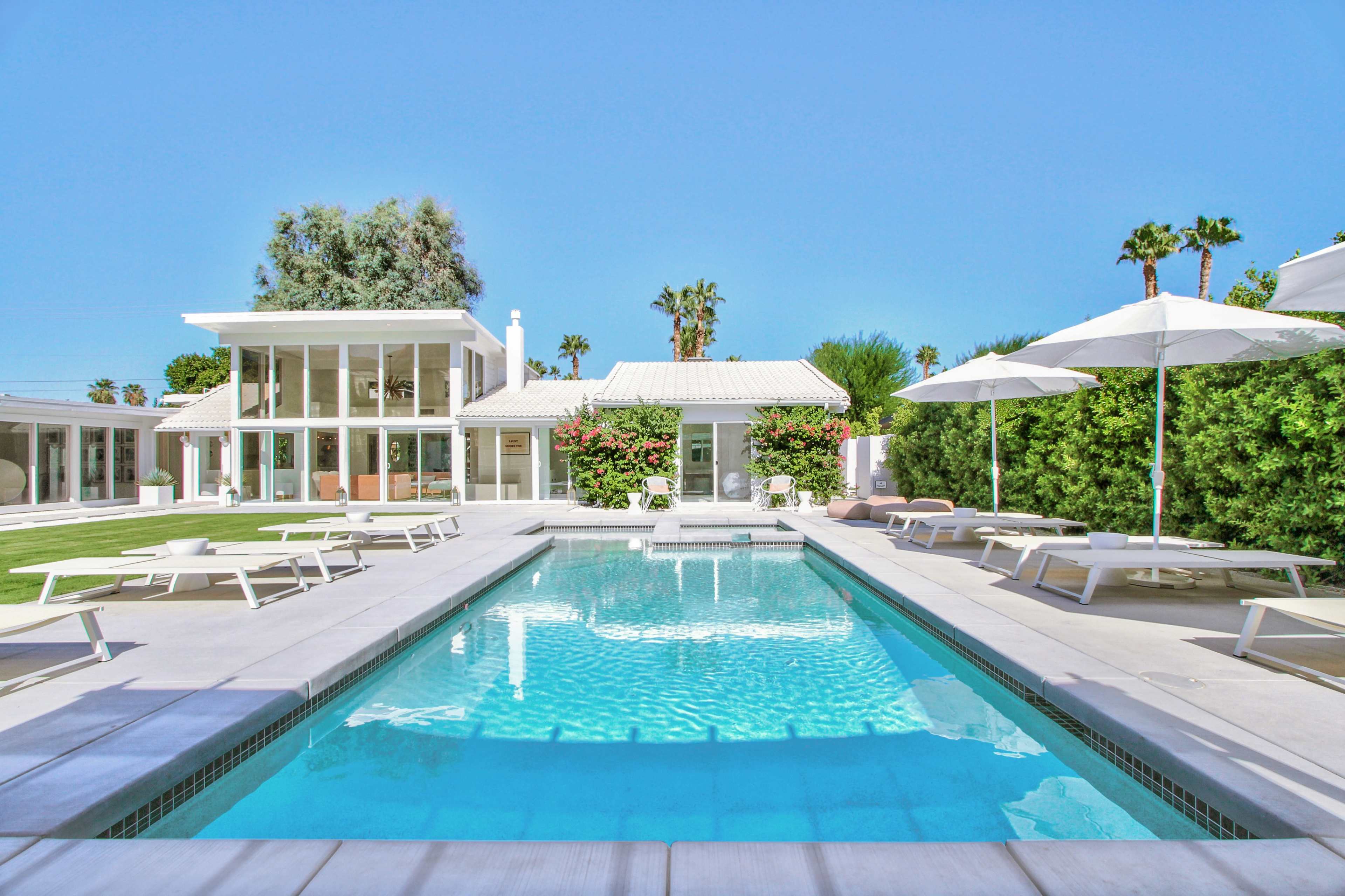 The image features a modern pool area surrounded by lounge chairs, with a clear blue pool and a white house in the background, complemented by palm trees and landscaped greenery.