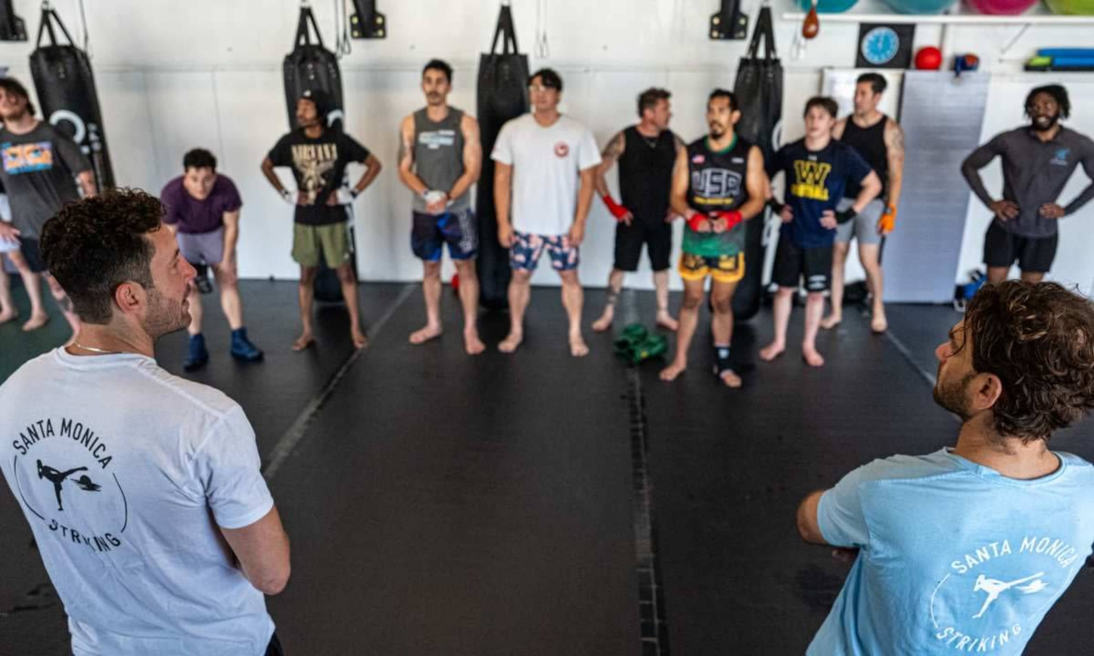 A group of individuals stands in a training area, facing two instructors at the front, surrounded by punching bags and exercise equipment.