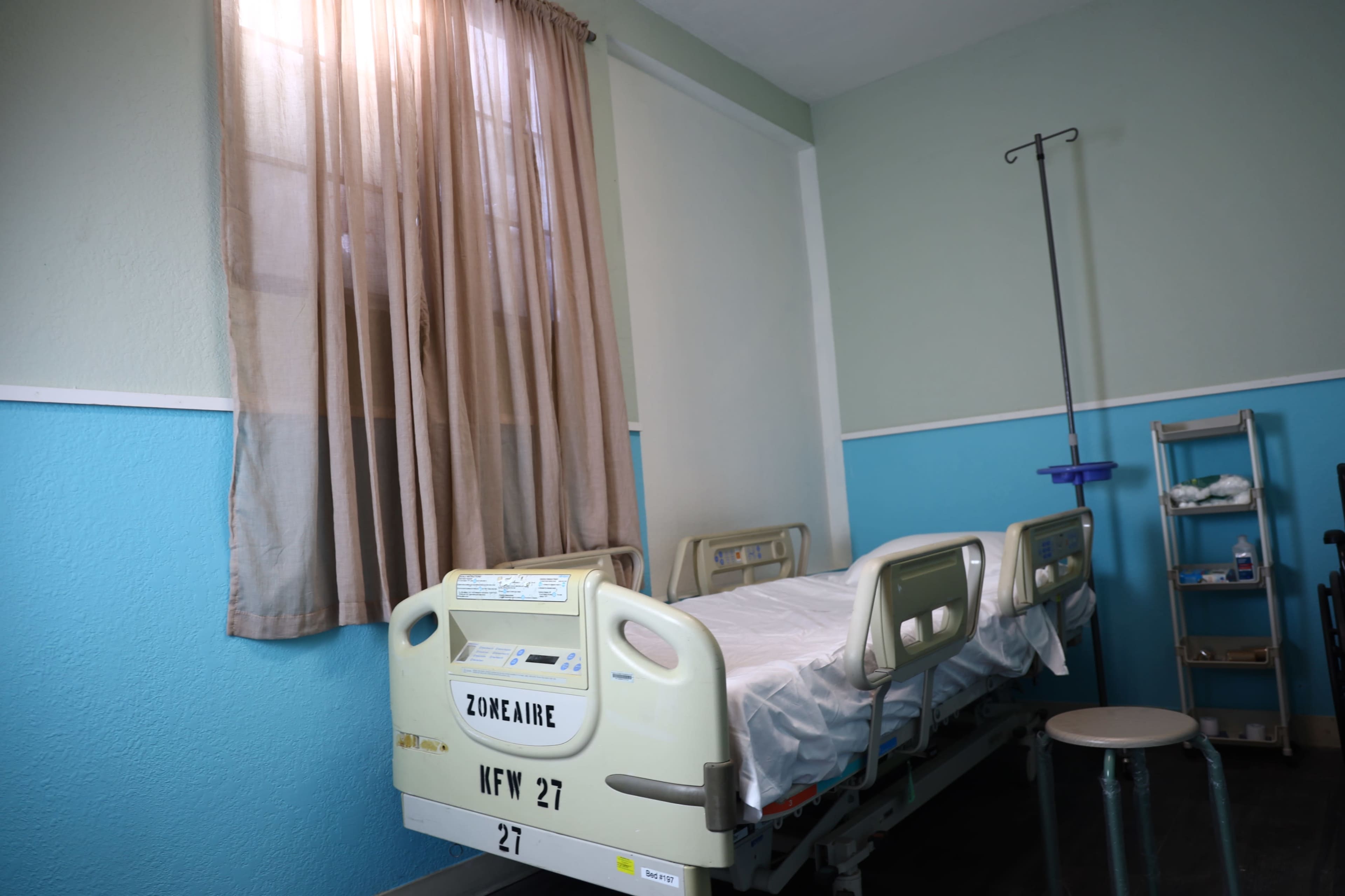 The image shows a hospital room with a single bed, a rolling IV stand, and a window covered with light curtains.