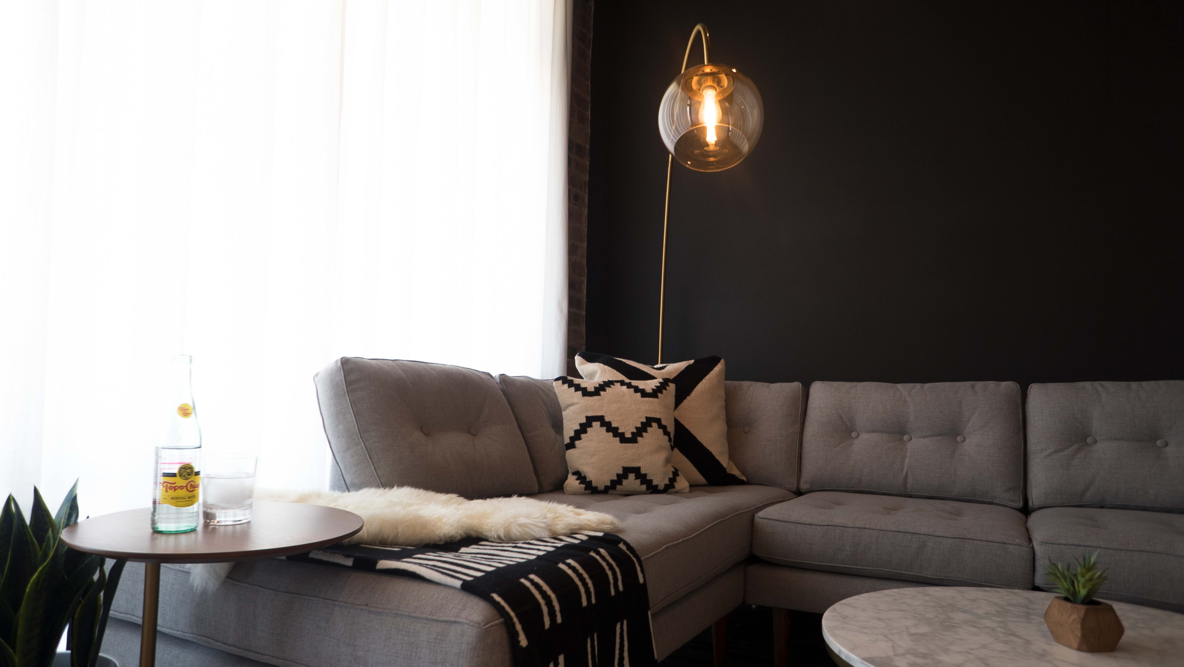 A modern living room features a gray sectional couch adorned with patterned cushions, a side table holding a drink and glass, and a sleek pendant light against a dark wall.