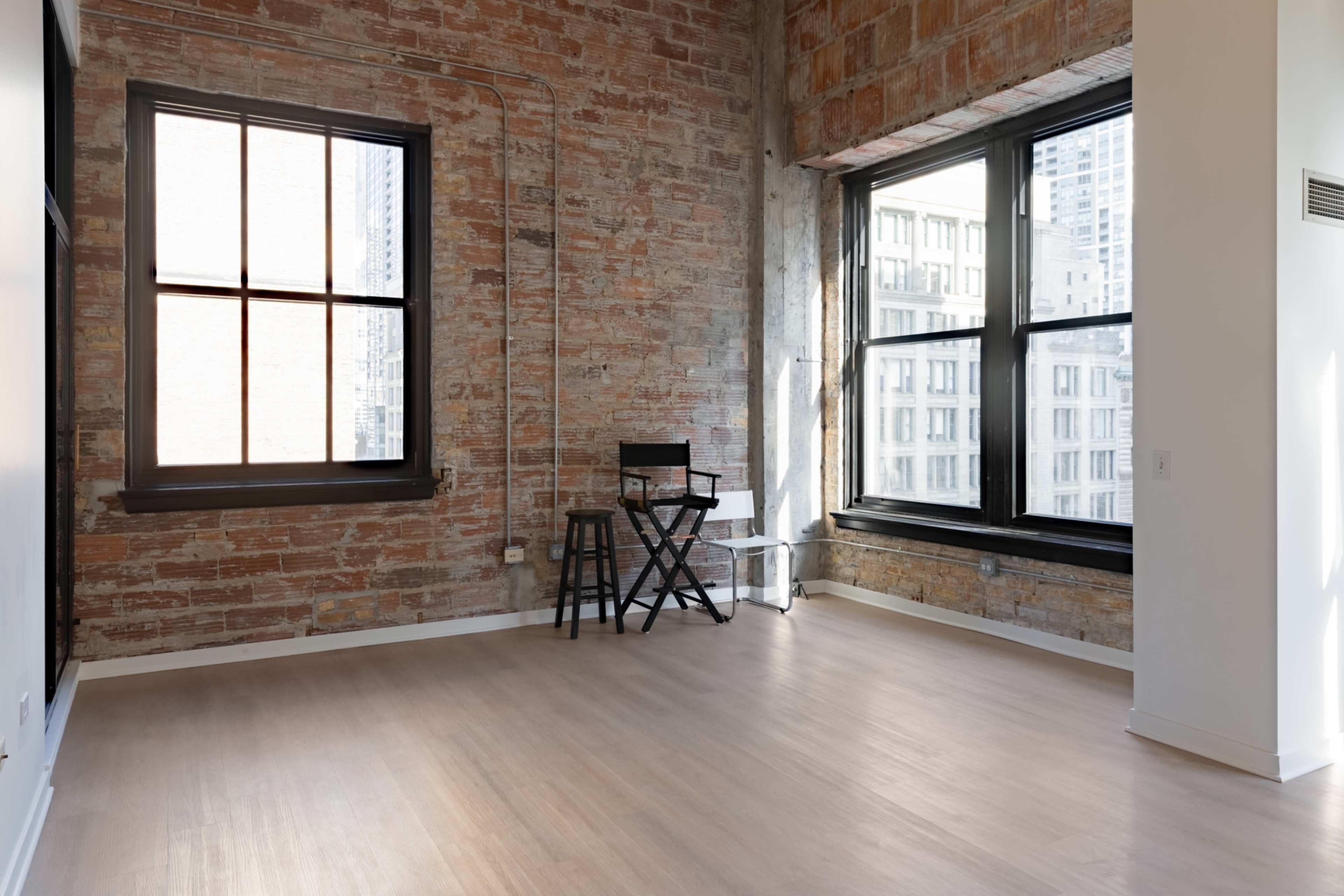 The image shows a sparsely furnished room with exposed brick walls and large windows allowing natural light to enter.