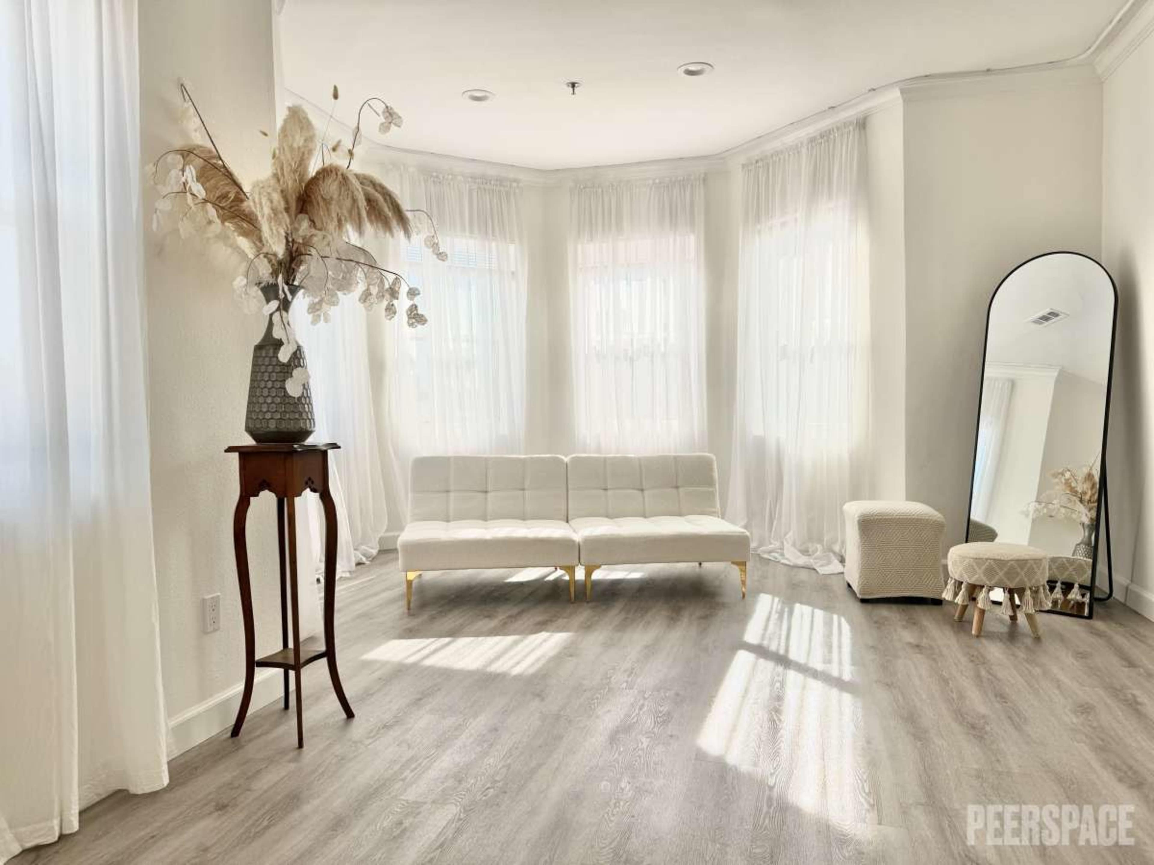 The image shows a bright, minimalist room featuring a white sofa, a round mirror, and a decorative plant arrangement on a small table.
