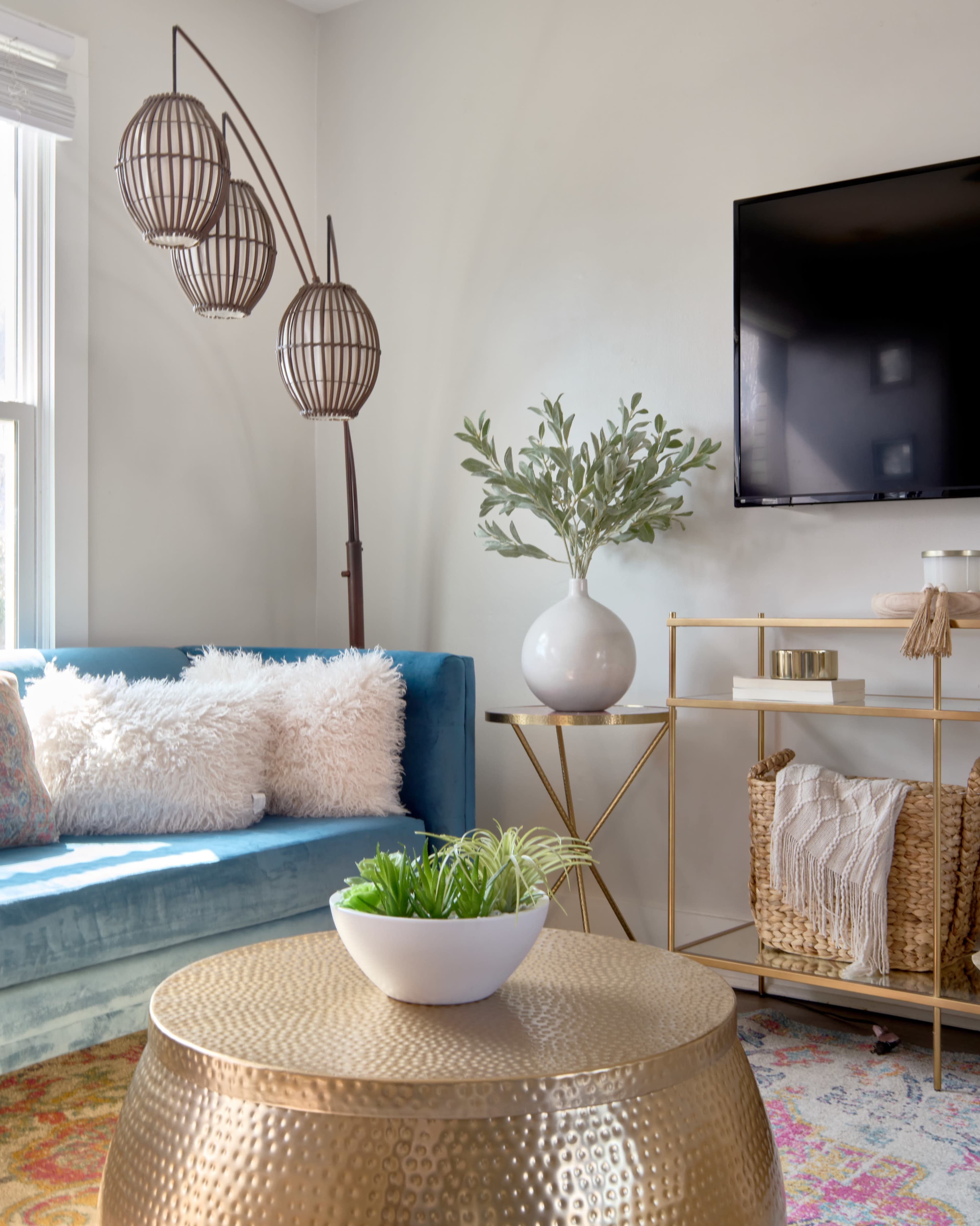 The image shows a modern living room with a blue couch, a gold coffee table, a decorative plant in a white bowl, and a flat-screen TV mounted on the wall.