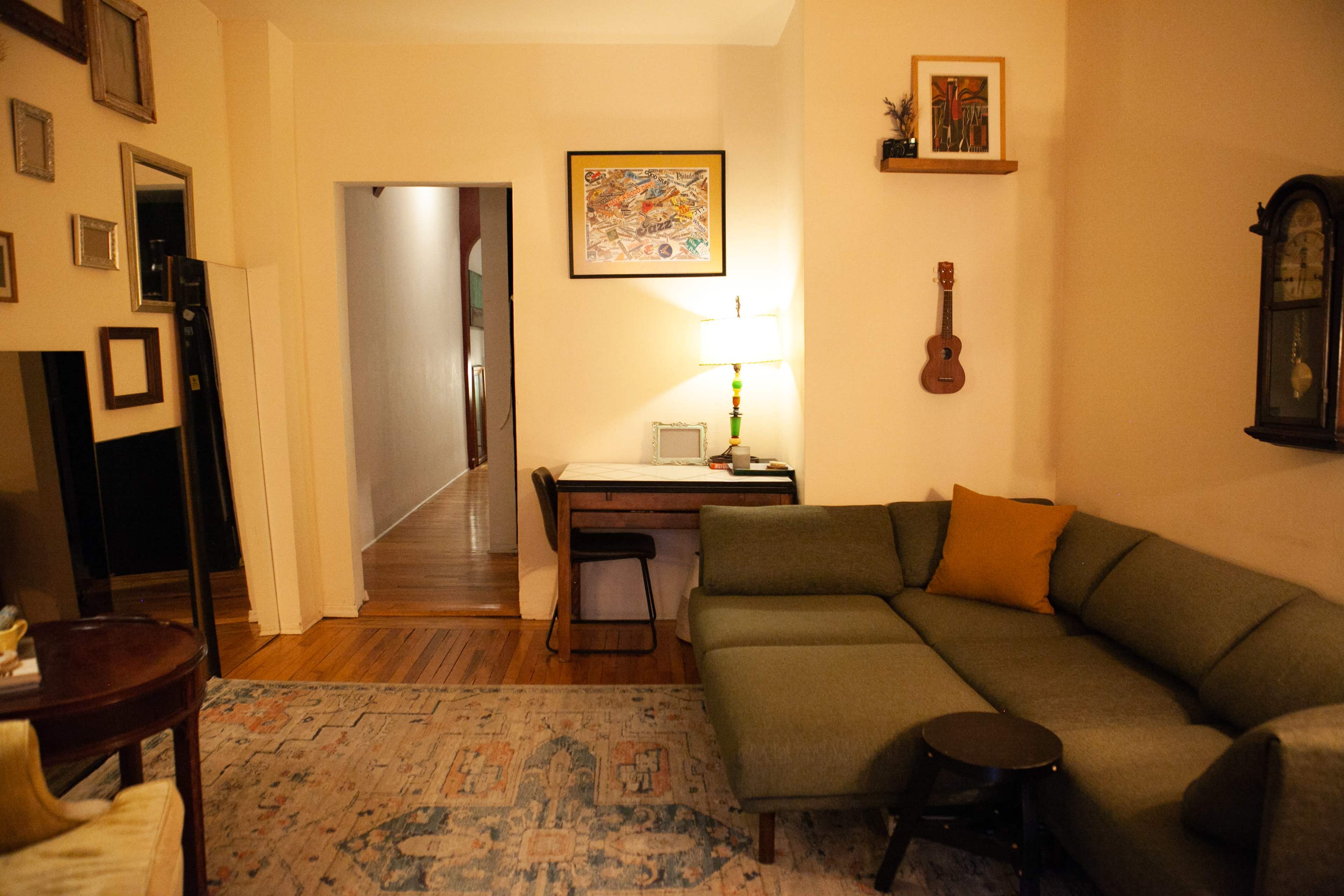 The scene depicts a cozy living room with a green sectional sofa, a small wooden side table, a desk with a chair, and various framed art pieces on the walls.