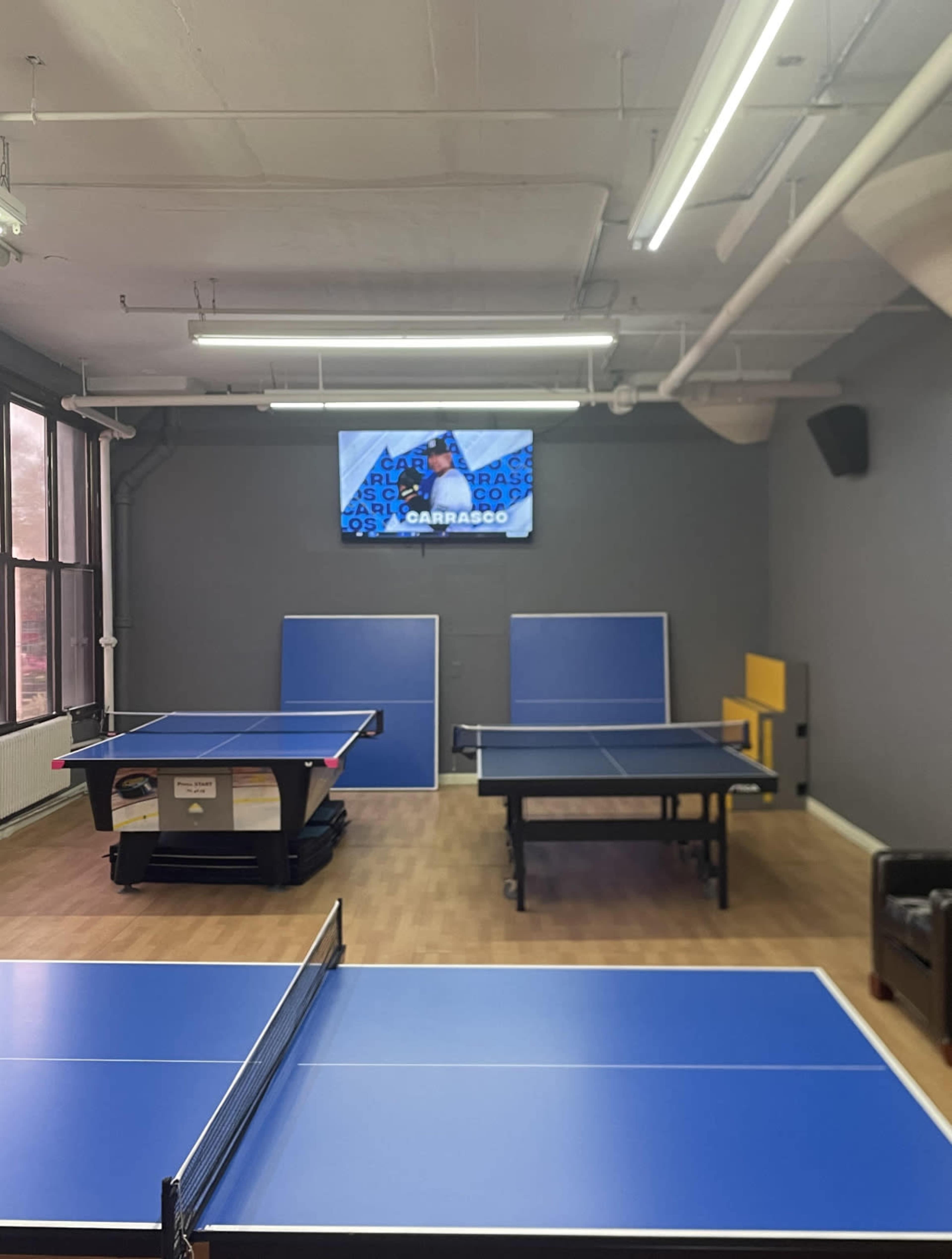The image shows a room with two ping pong tables, a yellow bench, and a wall-mounted screen displaying a player.
