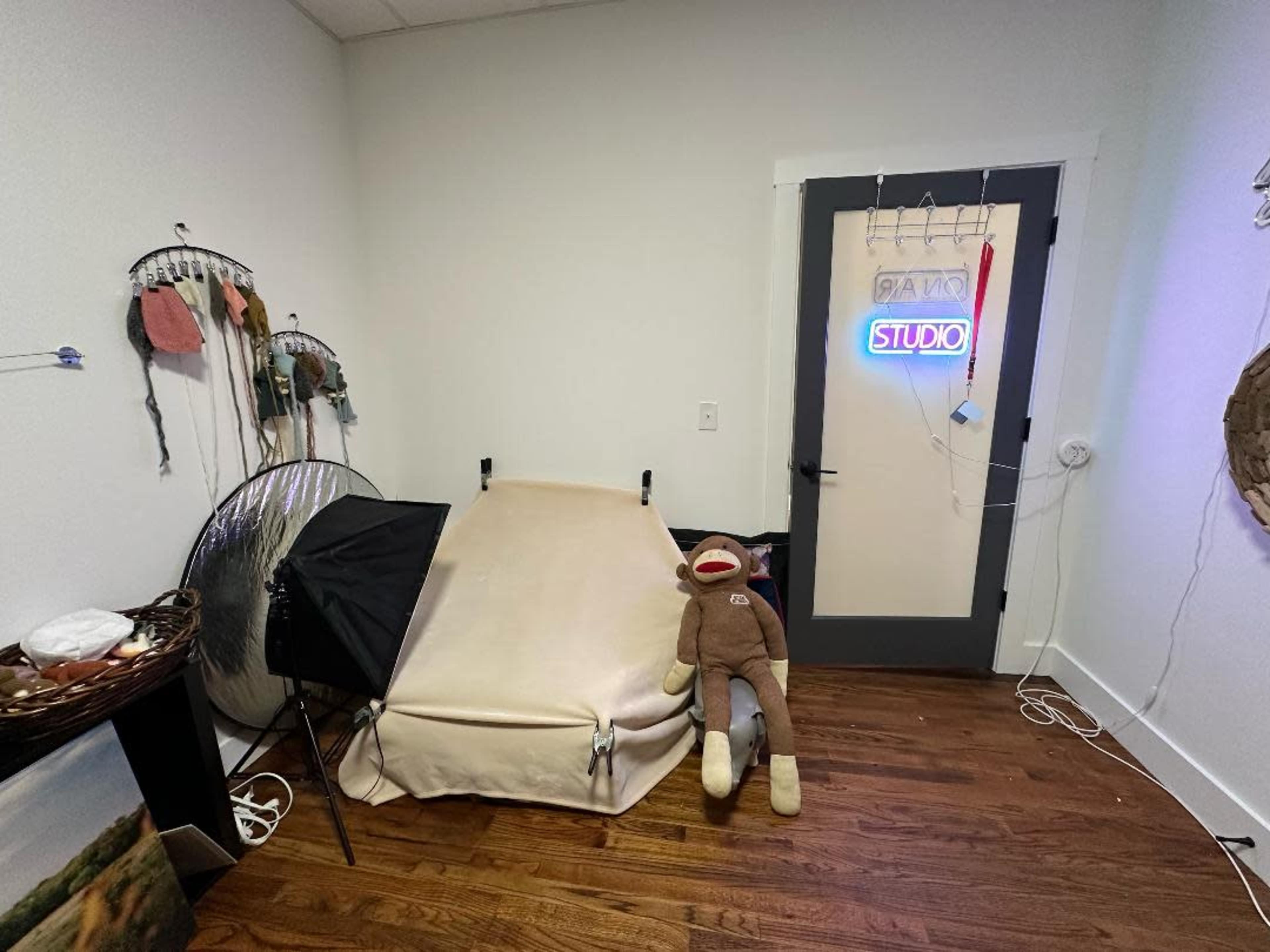 A small room with a light-colored backdrop, a softbox light, a stuffed monkey sitting on the floor, and a door featuring a neon sign that reads "STUDIO."