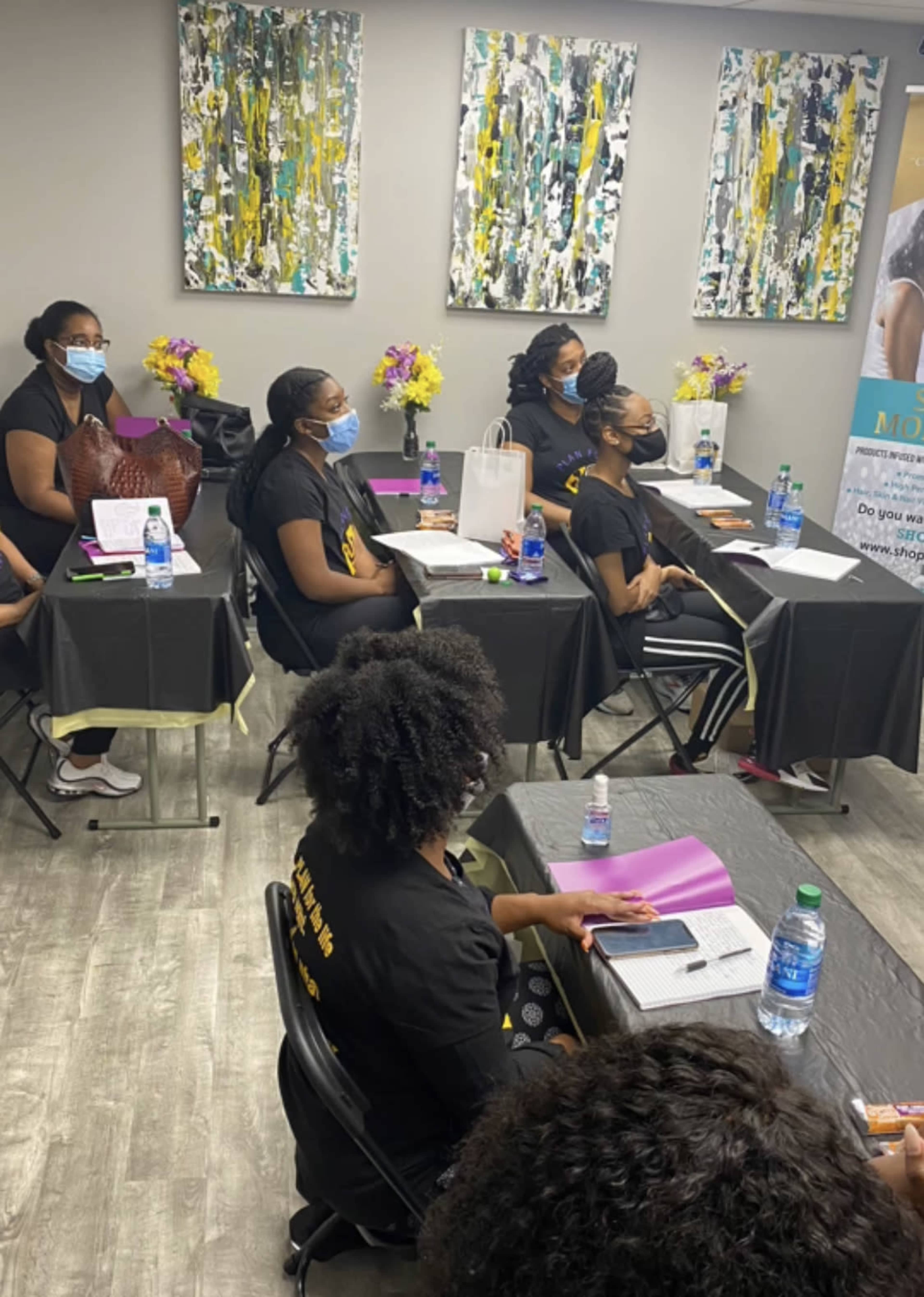 A group of women wearing black shirts and masks sit at tables in a classroom setting, focused on a presentation, with colorful artwork on the walls and flowers on the tables.