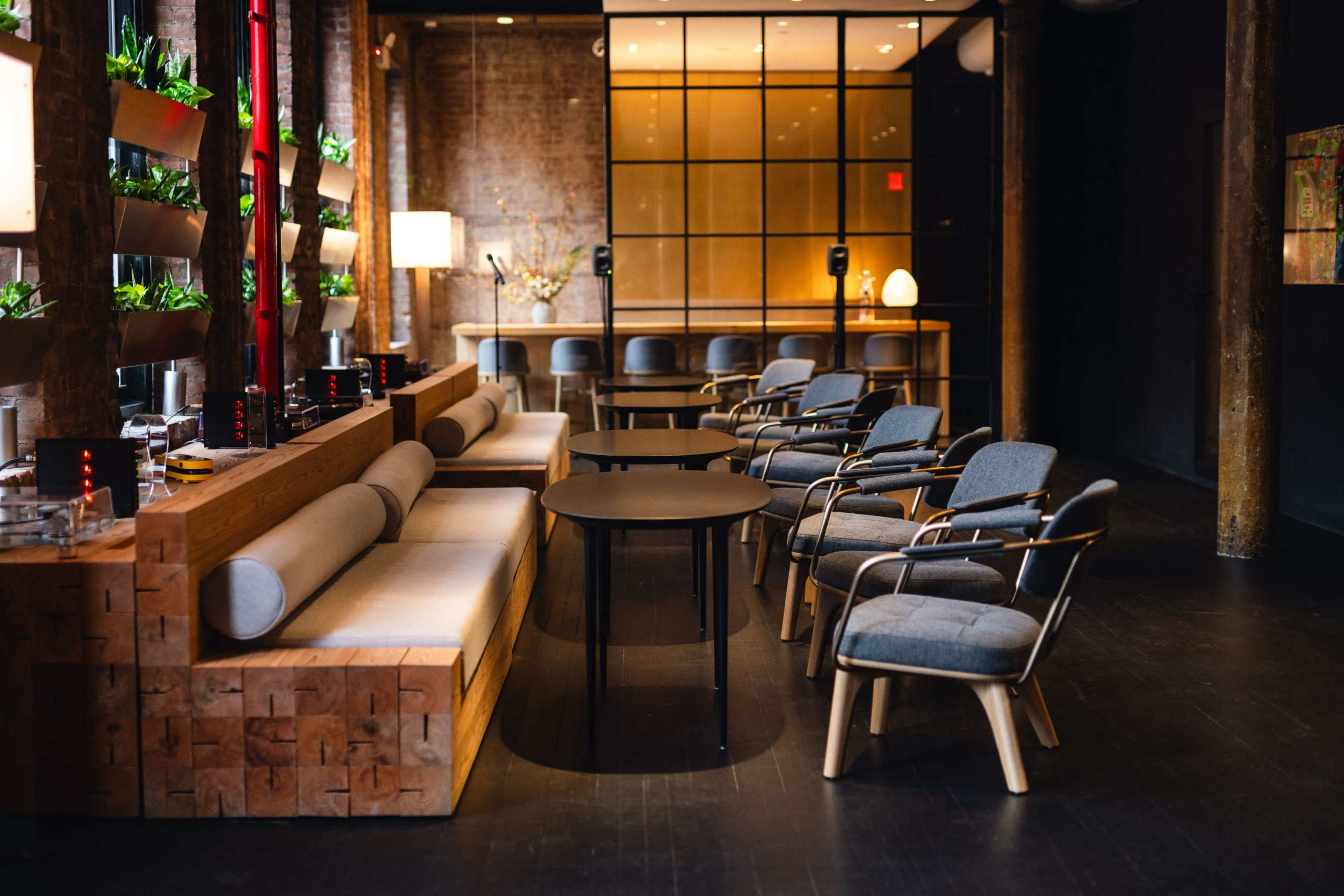 The image shows a modern lounge area with a long bench, several cushioned chairs, a round table, and plants on display along the walls.