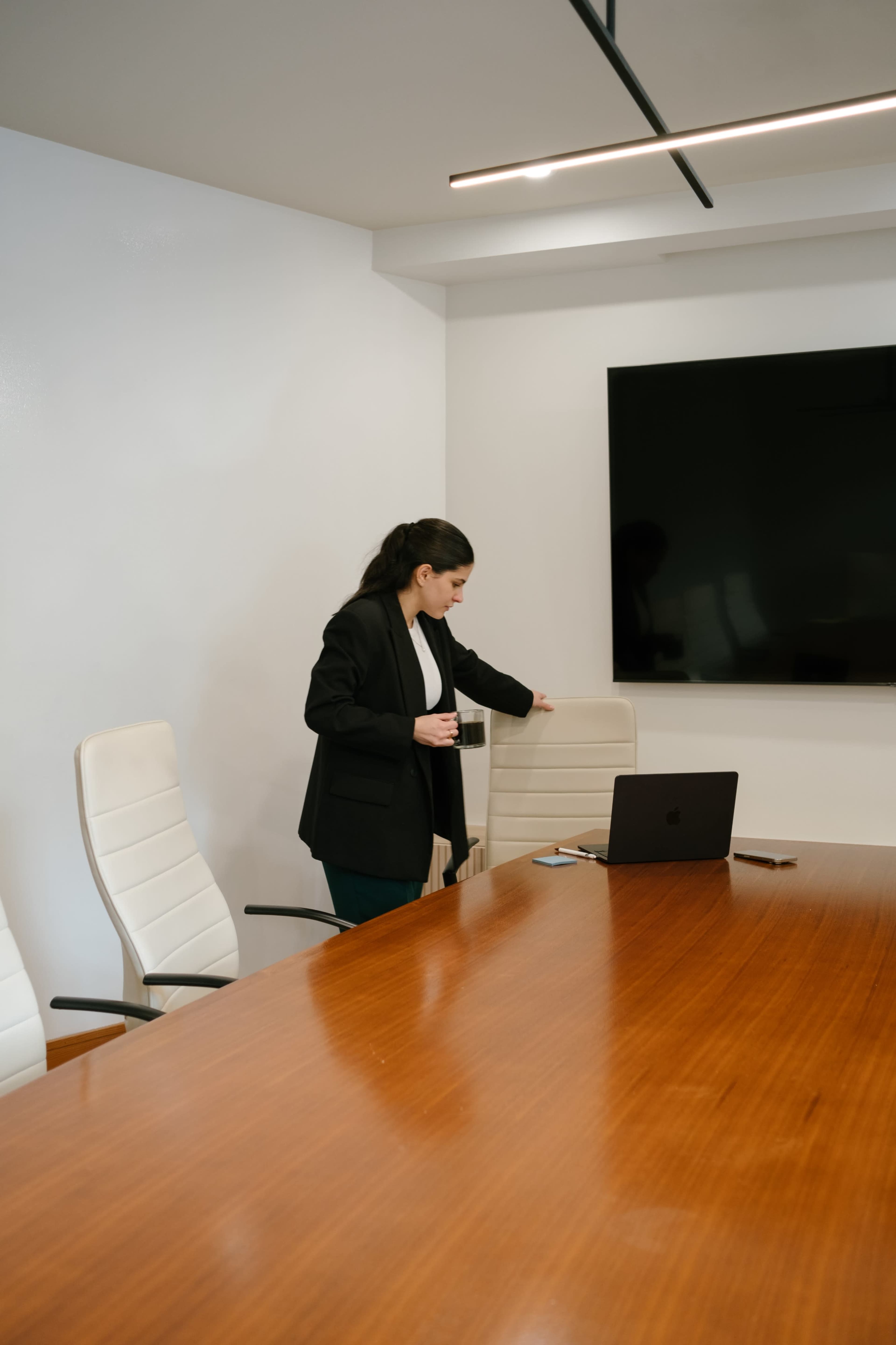 A woman in a black blazer adjusts a chair in a modern conference room with a large screen and a wooden table.