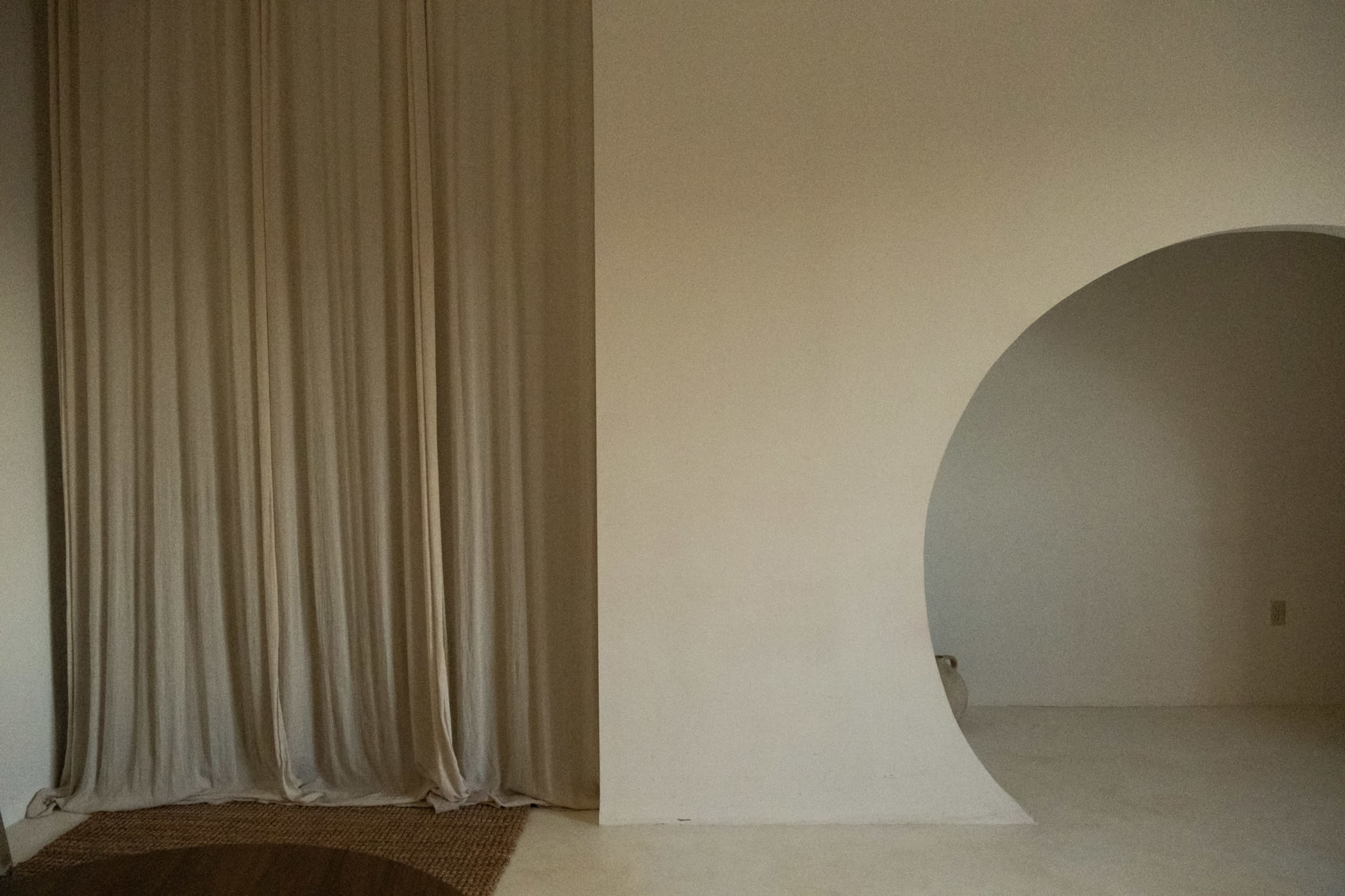 The image shows a minimalist interior space featuring a draped curtain on the left and a curved archway on the right against a plain wall.