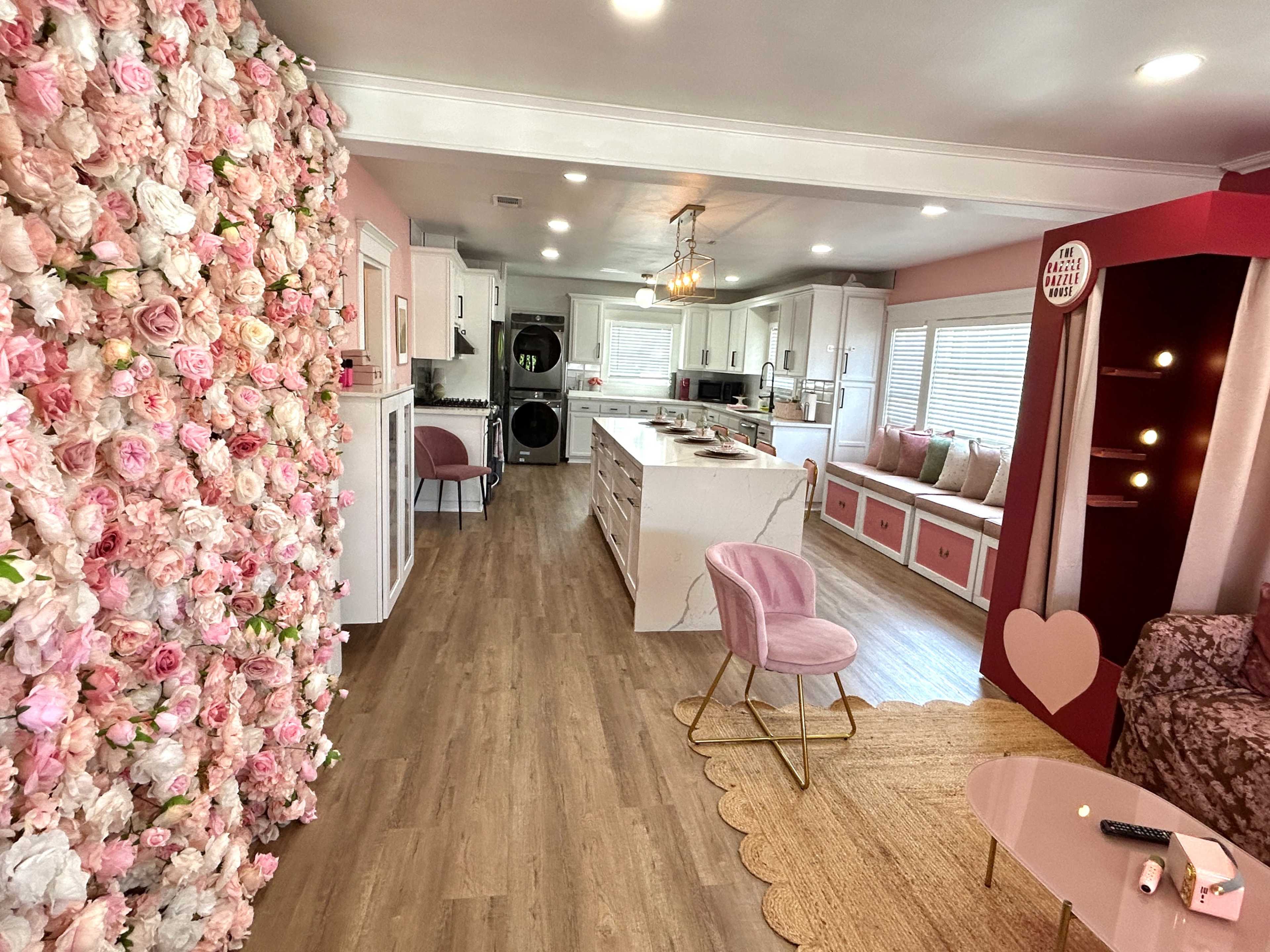 The image shows a brightly lit interior of a modern kitchen and living area, featuring a floral wall, pink accents, and stylish furnishings.