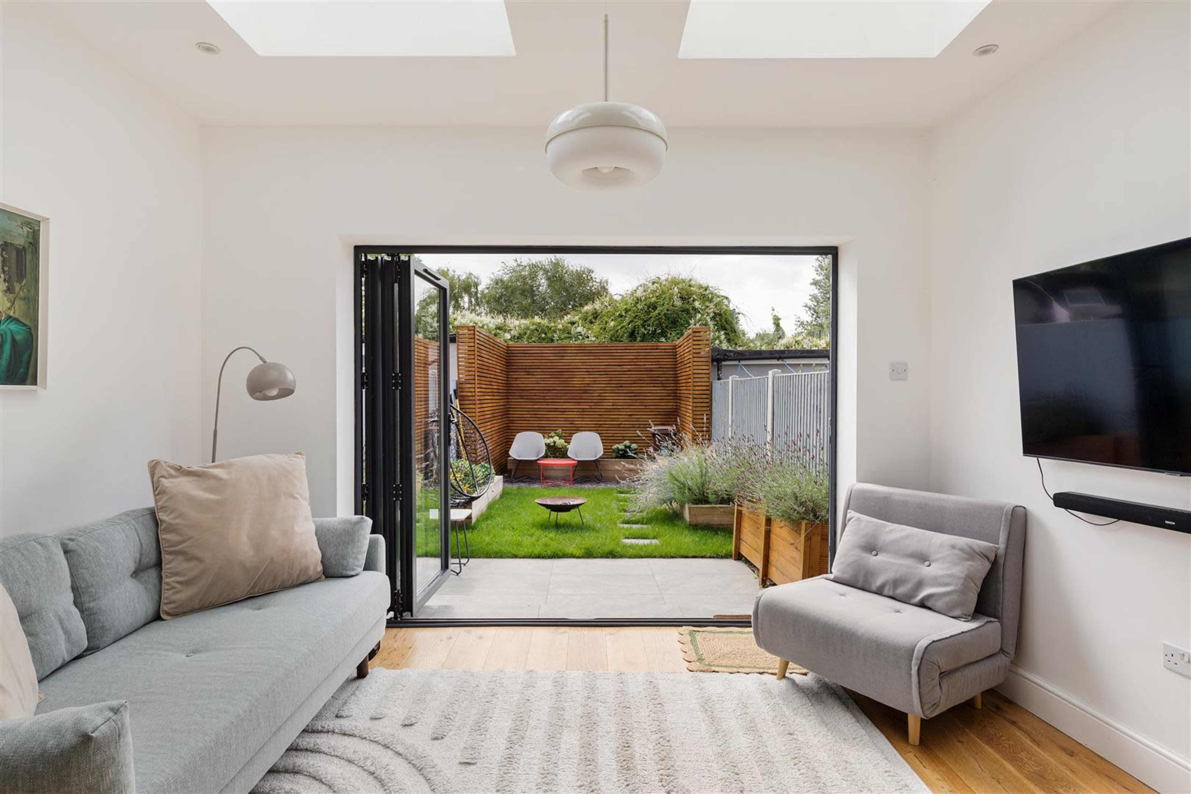 A modern living room features a gray sofa and a chair, with large open doors leading to a garden area adorned with greenery and wooden fencing.