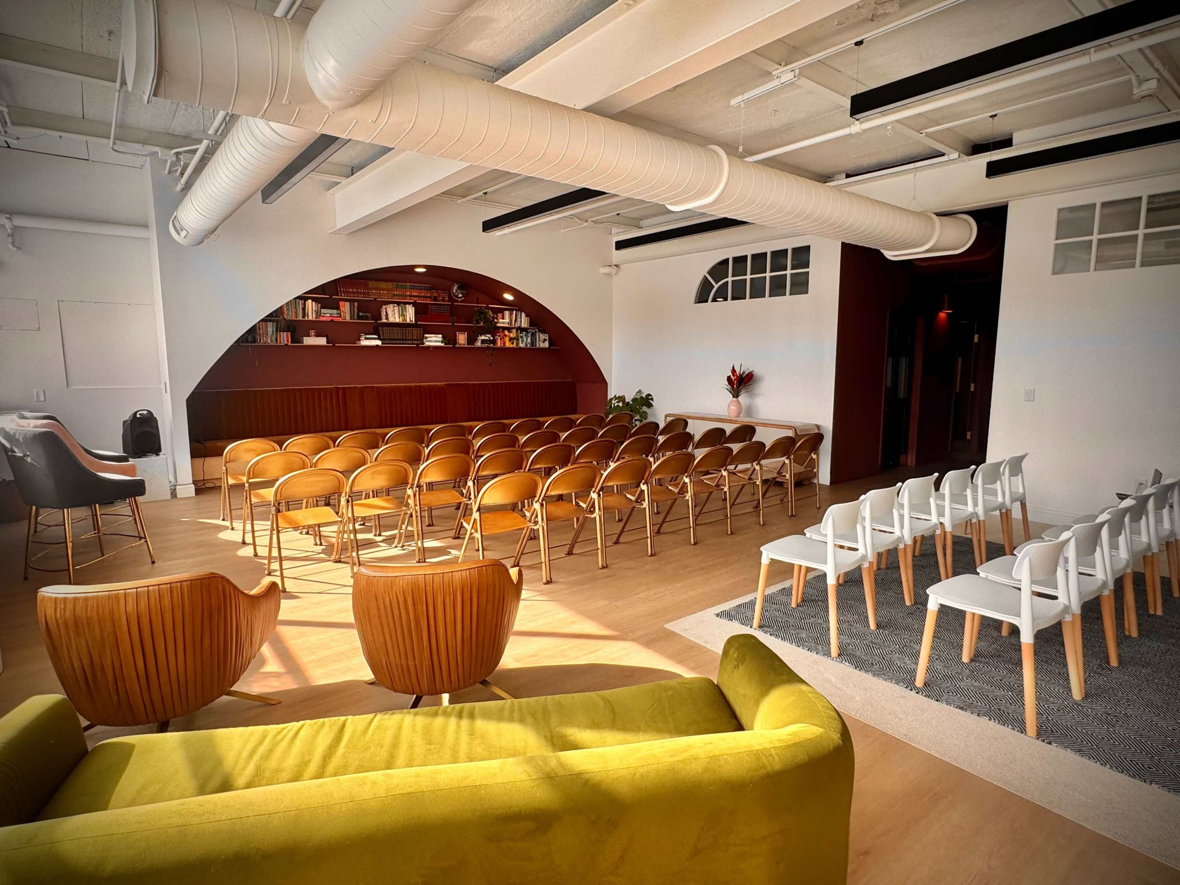 The image shows a modern event space with rows of wooden chairs and white chairs arranged for an audience, along with a green sofa in the foreground and shelves in the background.