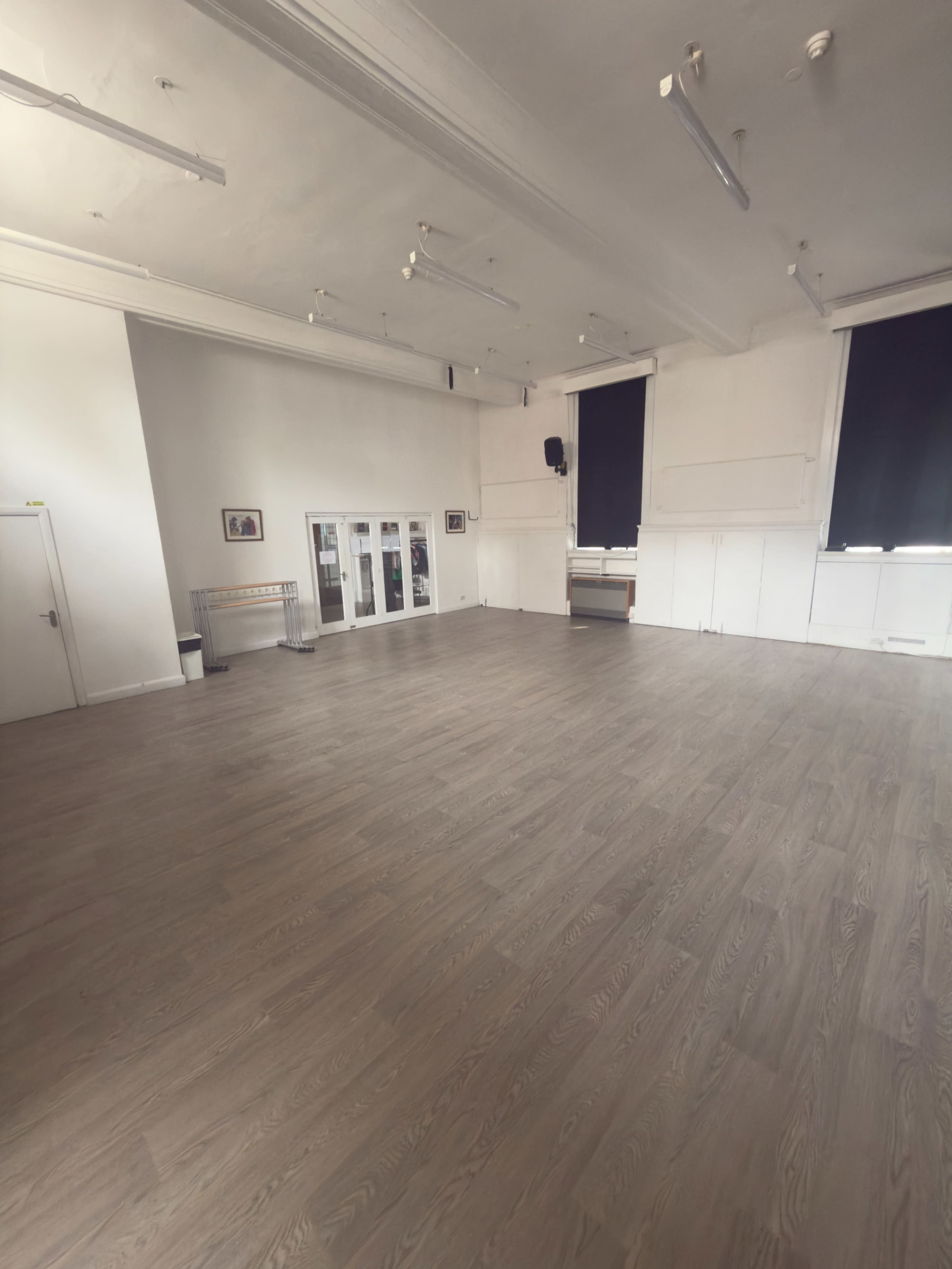 The image shows a spacious, empty room with wooden flooring, large windows with black coverings, and white walls.