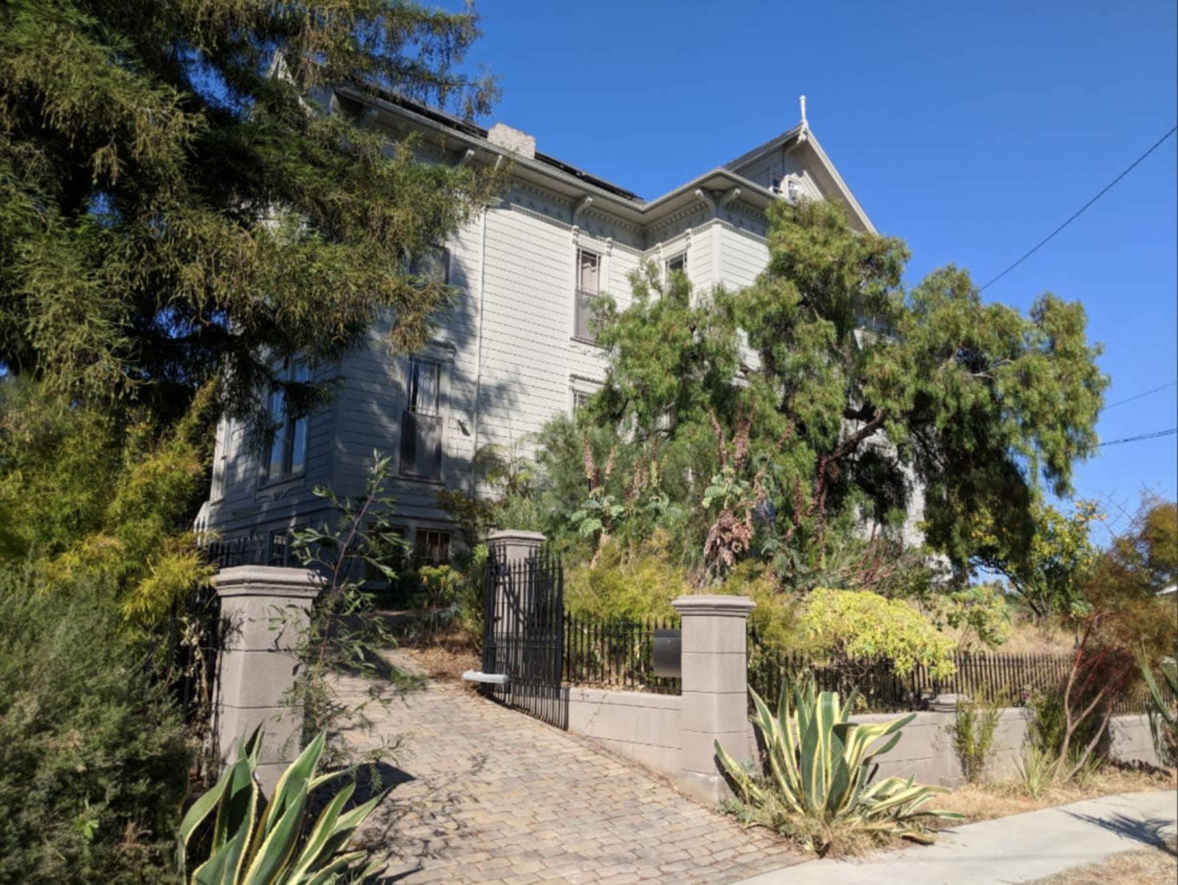A large, two-story gray house with a steep roof is partially obscured by overgrown trees and shrubs, accessible via a cobblestone driveway and surrounded by a low fence.