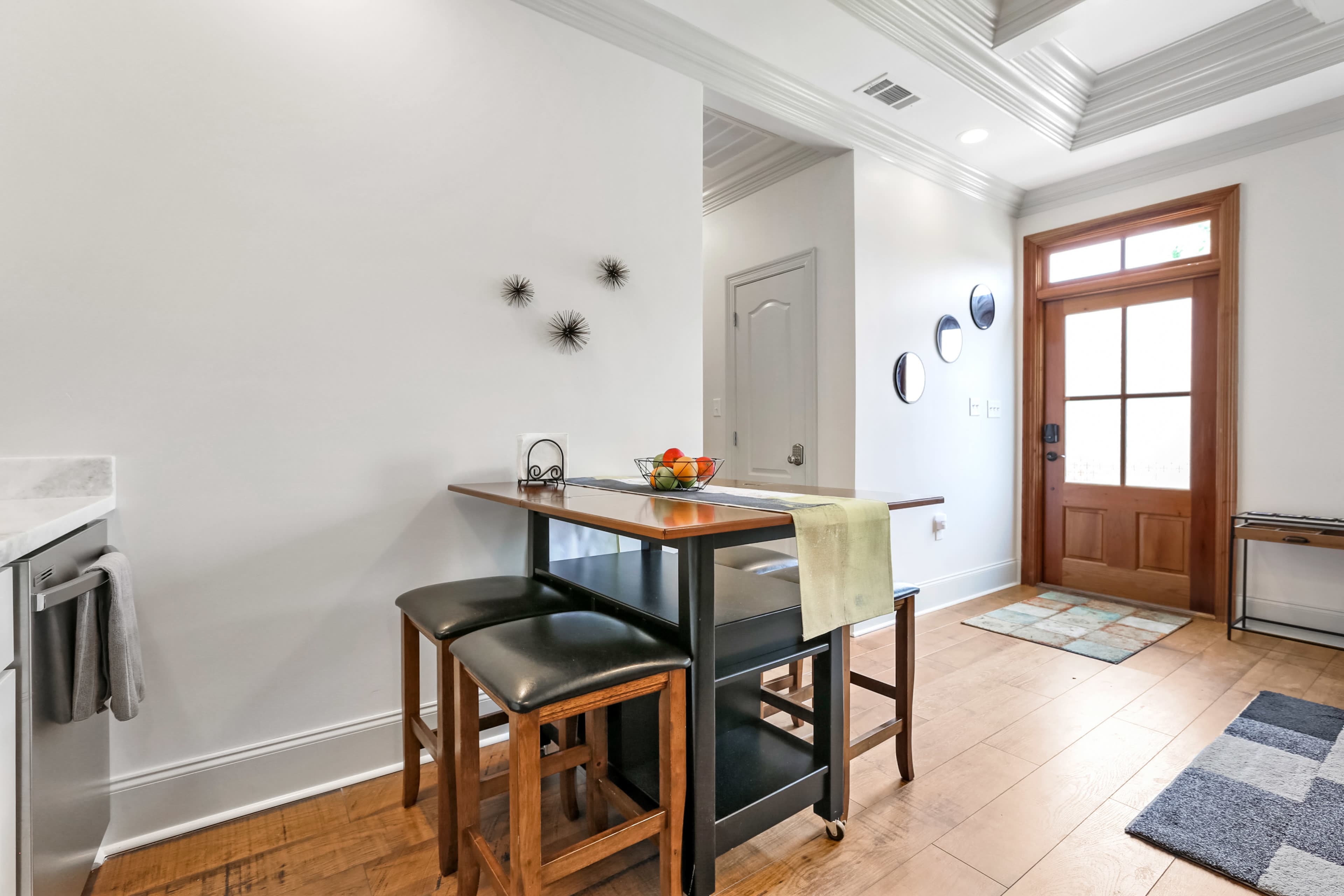 The image shows a modern kitchen area with a small table and two black stools, near a doorway with decorative circular wall art.