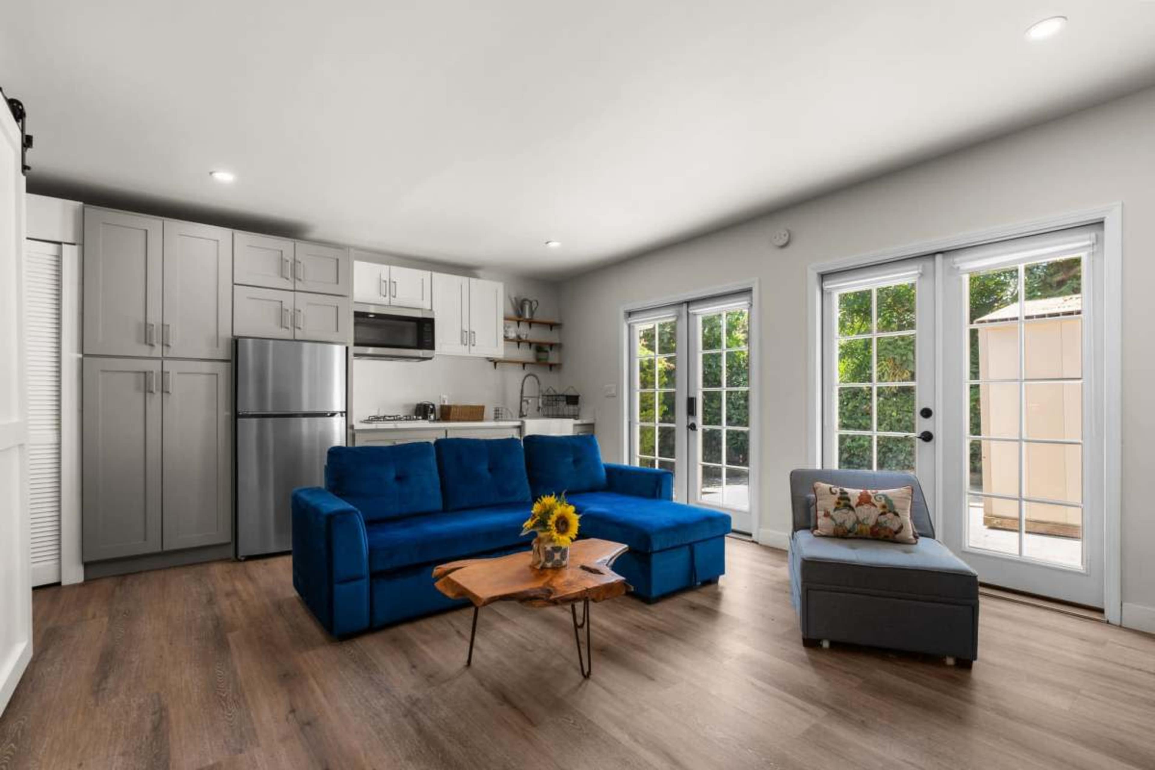 A small living area features a blue sofa, a wooden coffee table, and a kitchenette with stainless steel appliances, along with sliding glass doors leading to an outdoor space.