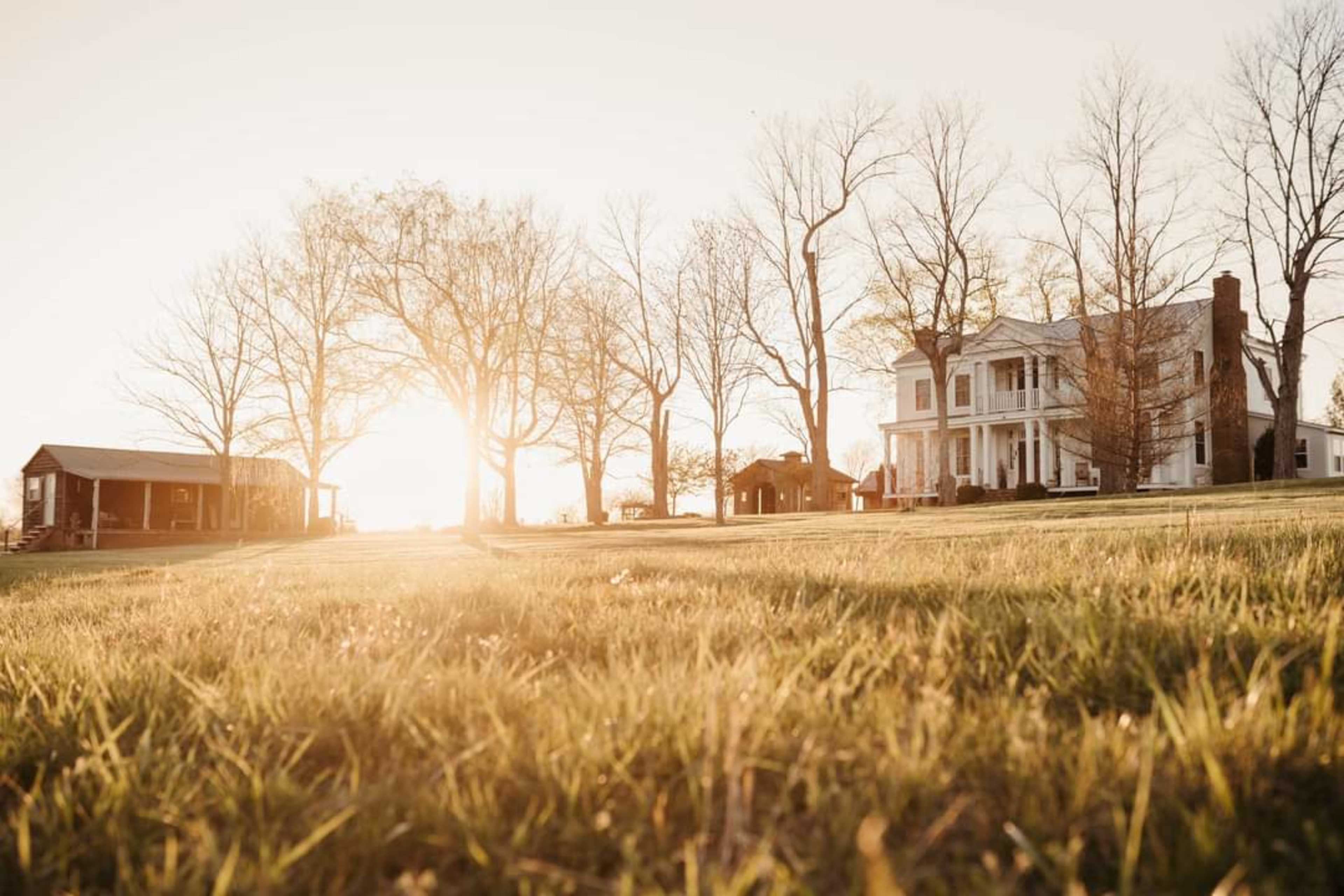 A large white house with a porch is set against the backdrop of a sunset, surrounded by bare trees and open grassy areas.