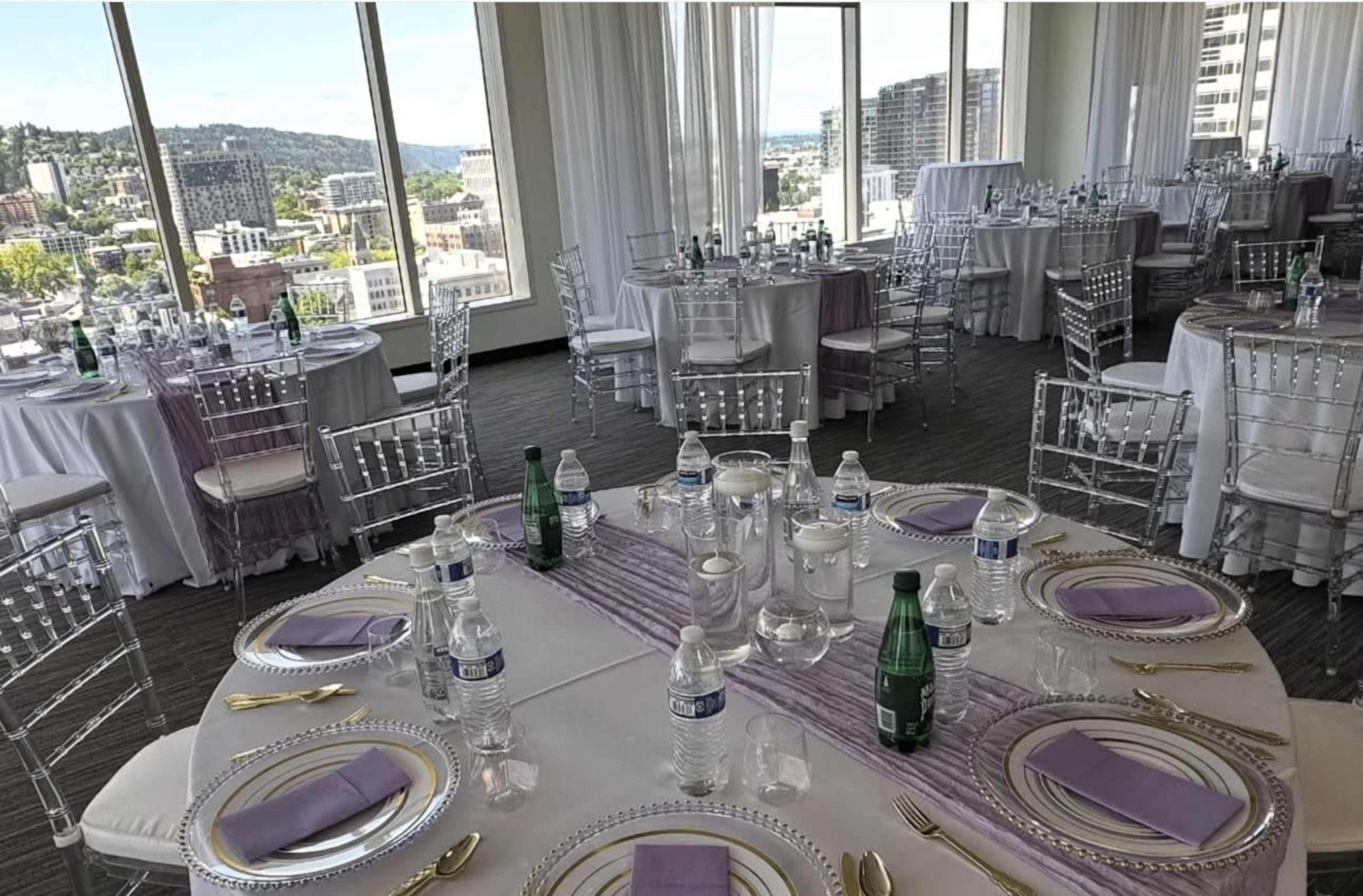 The image shows a banquet hall with round tables set for an event, featuring white tablecloths, clear chairs, and city views through large windows.