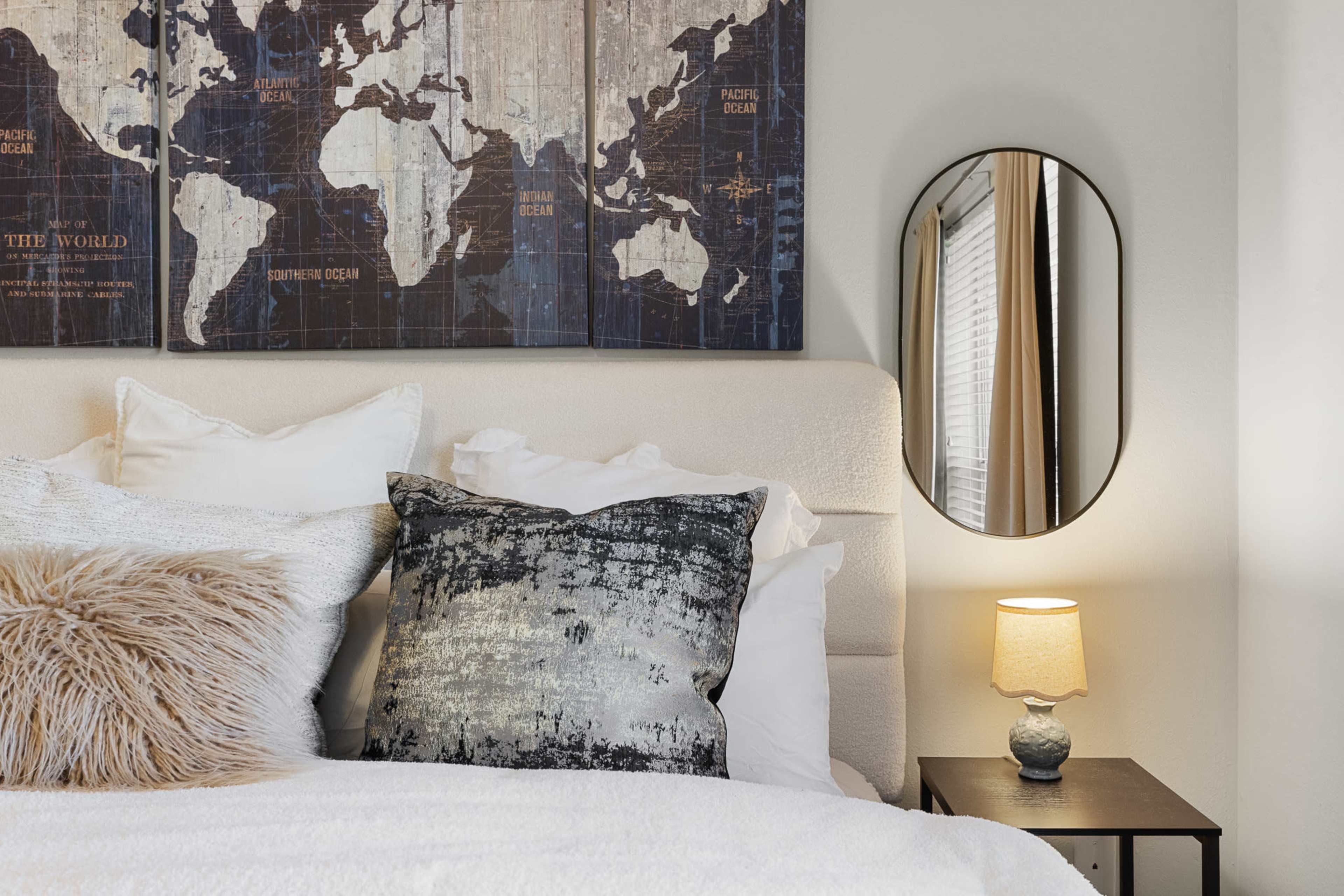 The image shows a neatly made bed with decorative pillows, an oval mirror, and a small bedside table with a lamp, all set against a wall featuring a world map.