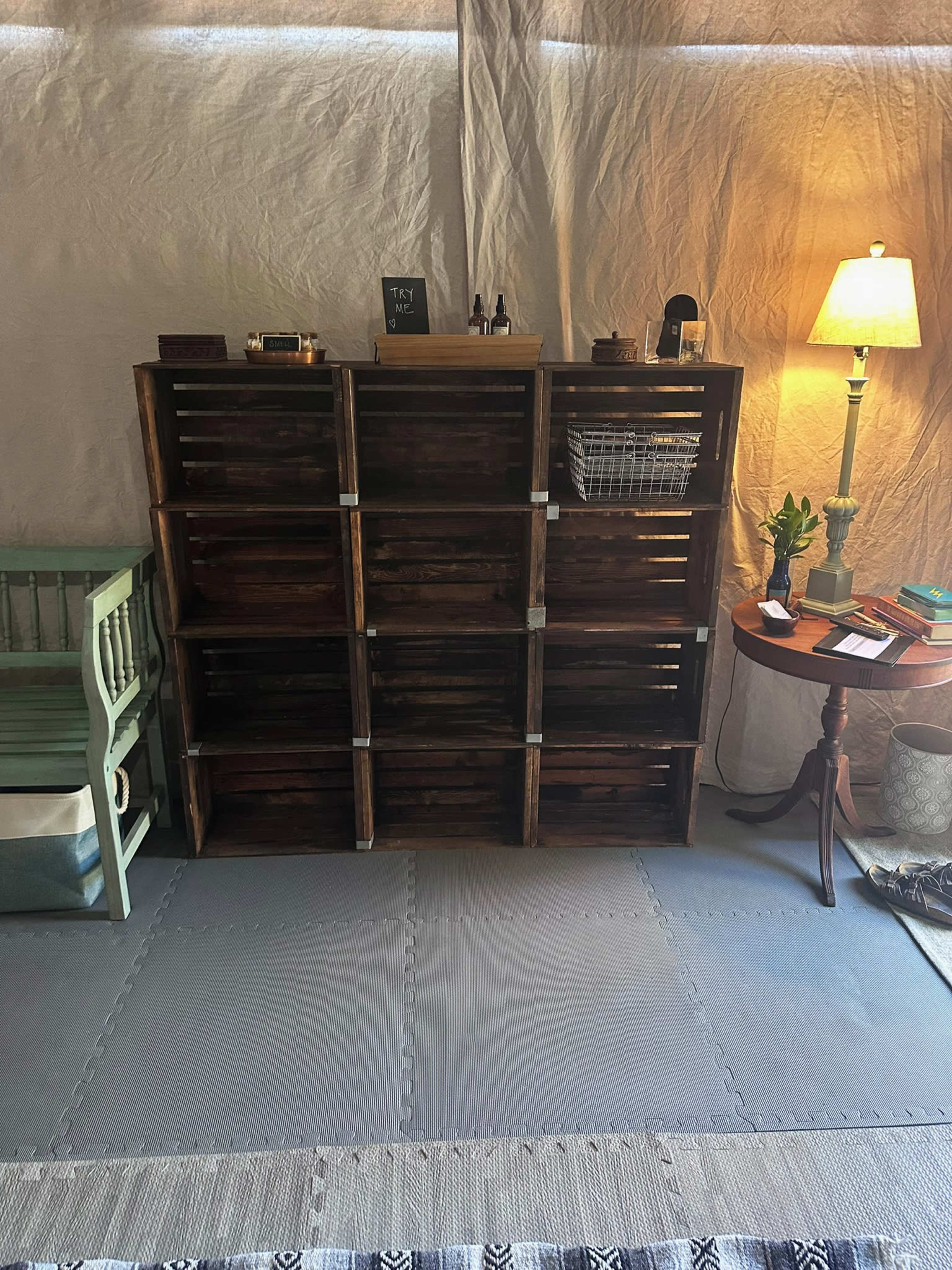 The image shows a wooden shelving unit with open crates, positioned next to a green chair and a small round table topped with a lamp and various items.
