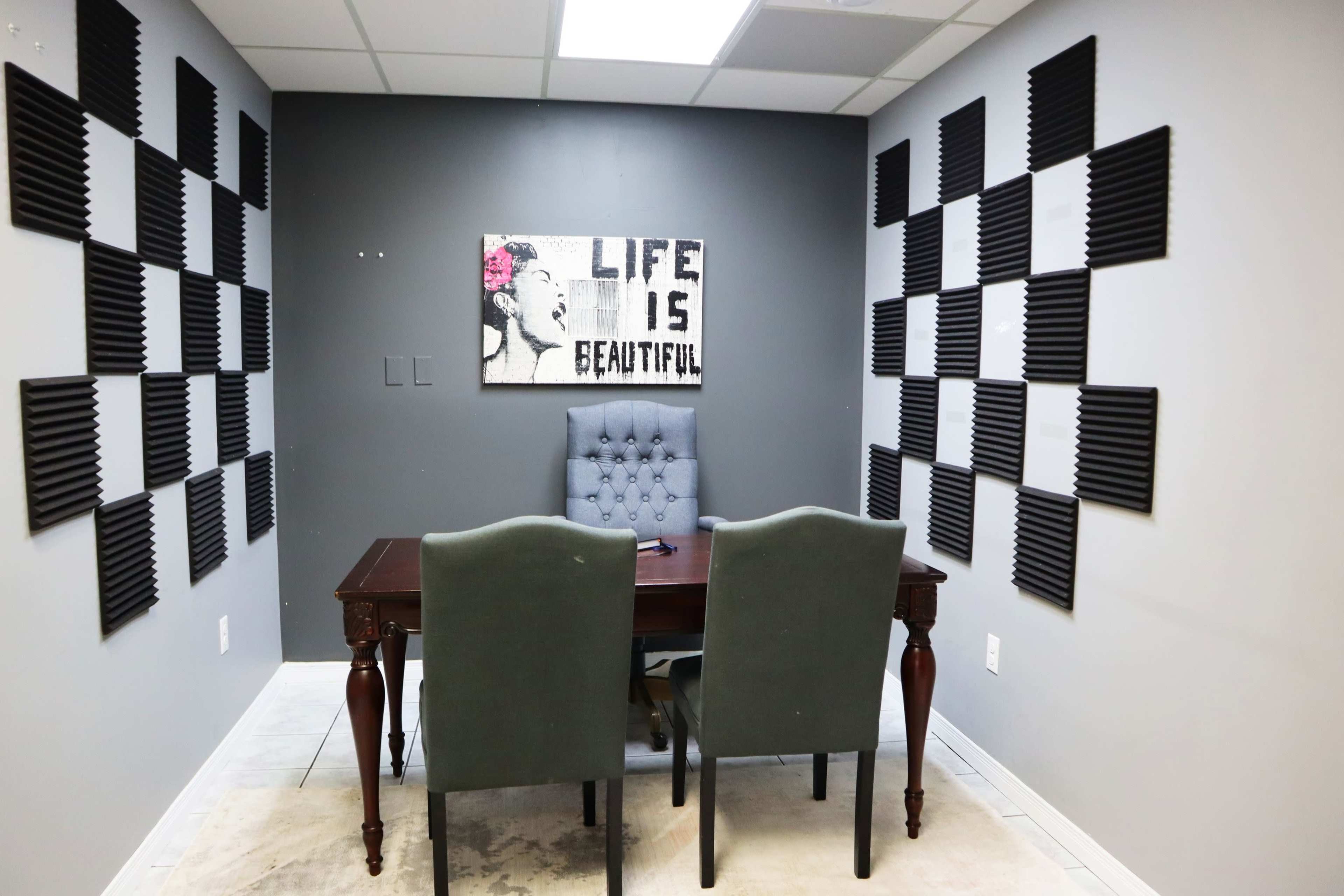 A small room features a table and two chairs, with soundproof panels on the walls and a framed artwork that reads "LIFE IS BEAUTIFUL."