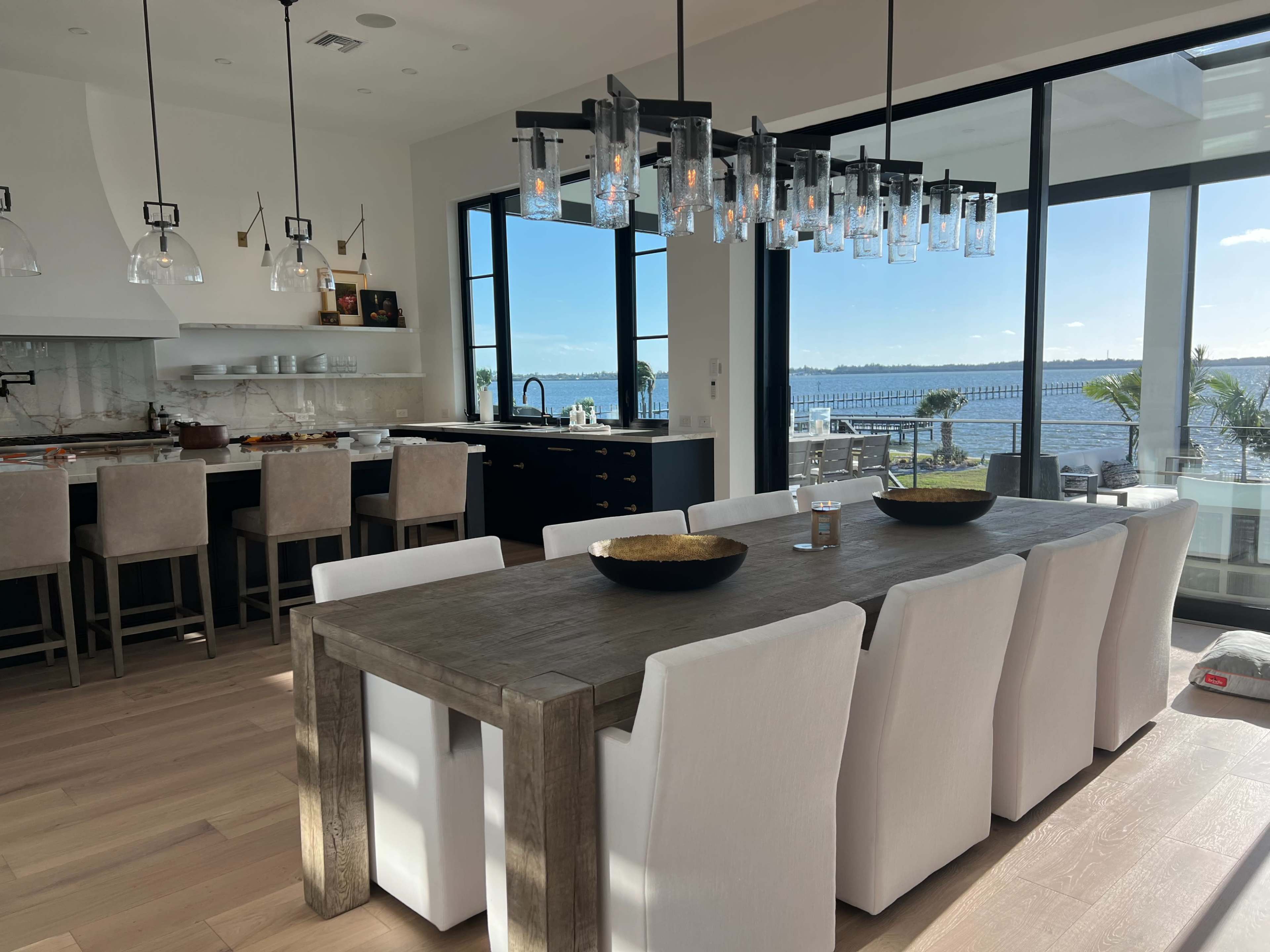 A modern dining area features a long wooden table surrounded by white upholstered chairs, with large windows showcasing a view of the water.