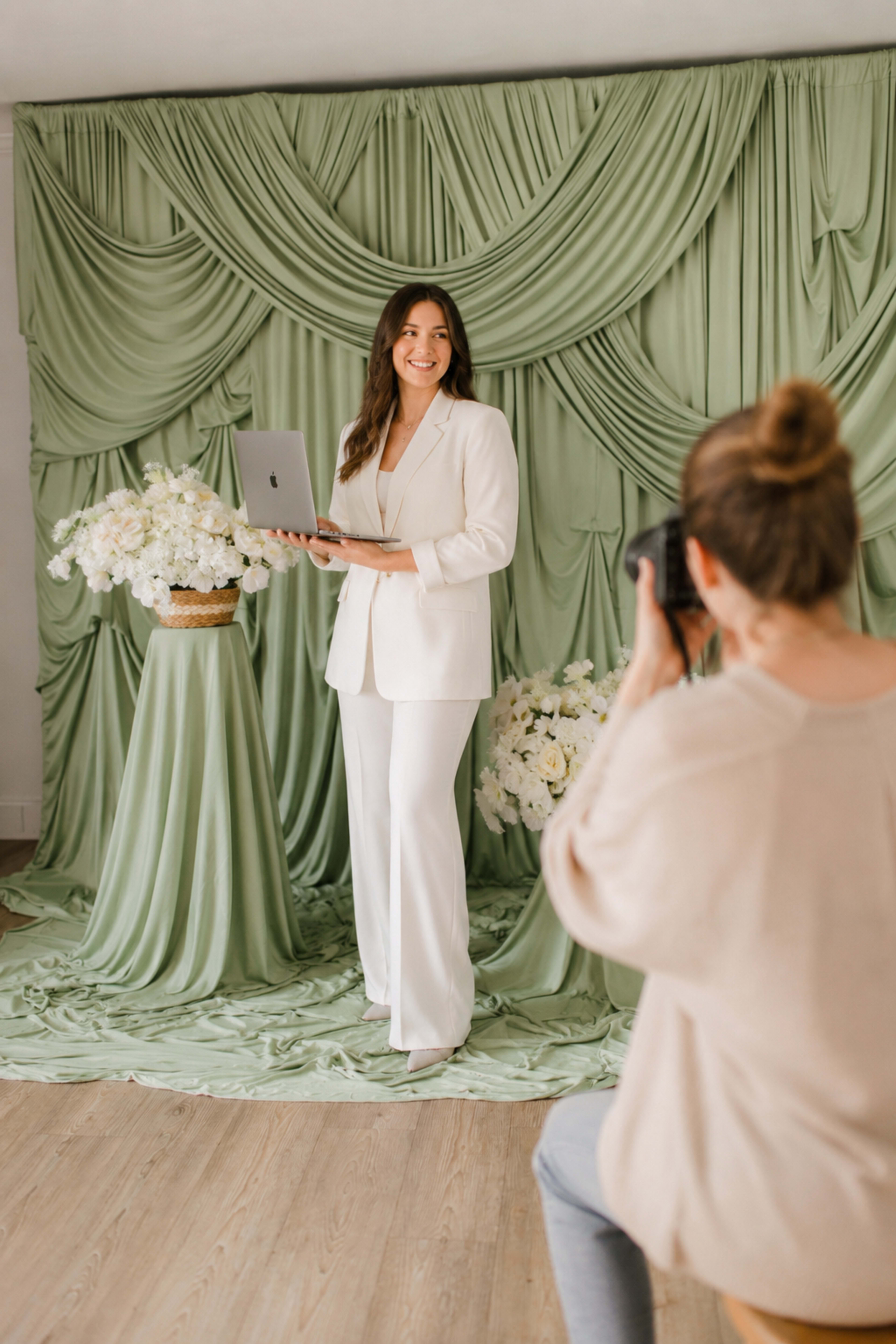 A woman in a white suit poses with a laptop in front of a green draped backdrop while another person takes her photograph.
