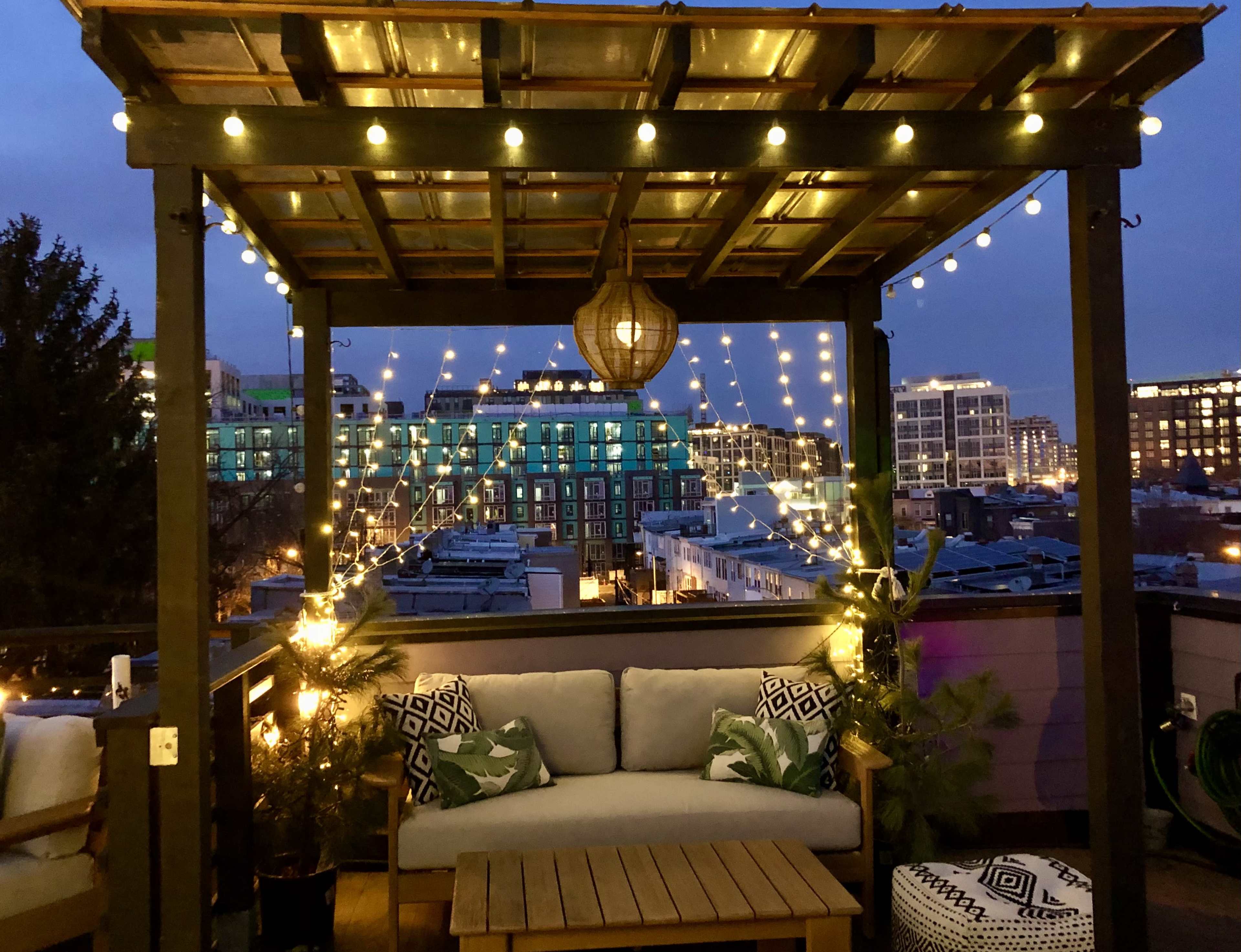 A rooftop seating area is adorned with string lights and plants, featuring a wooden table and cushions against a backdrop of city buildings under a twilight sky.