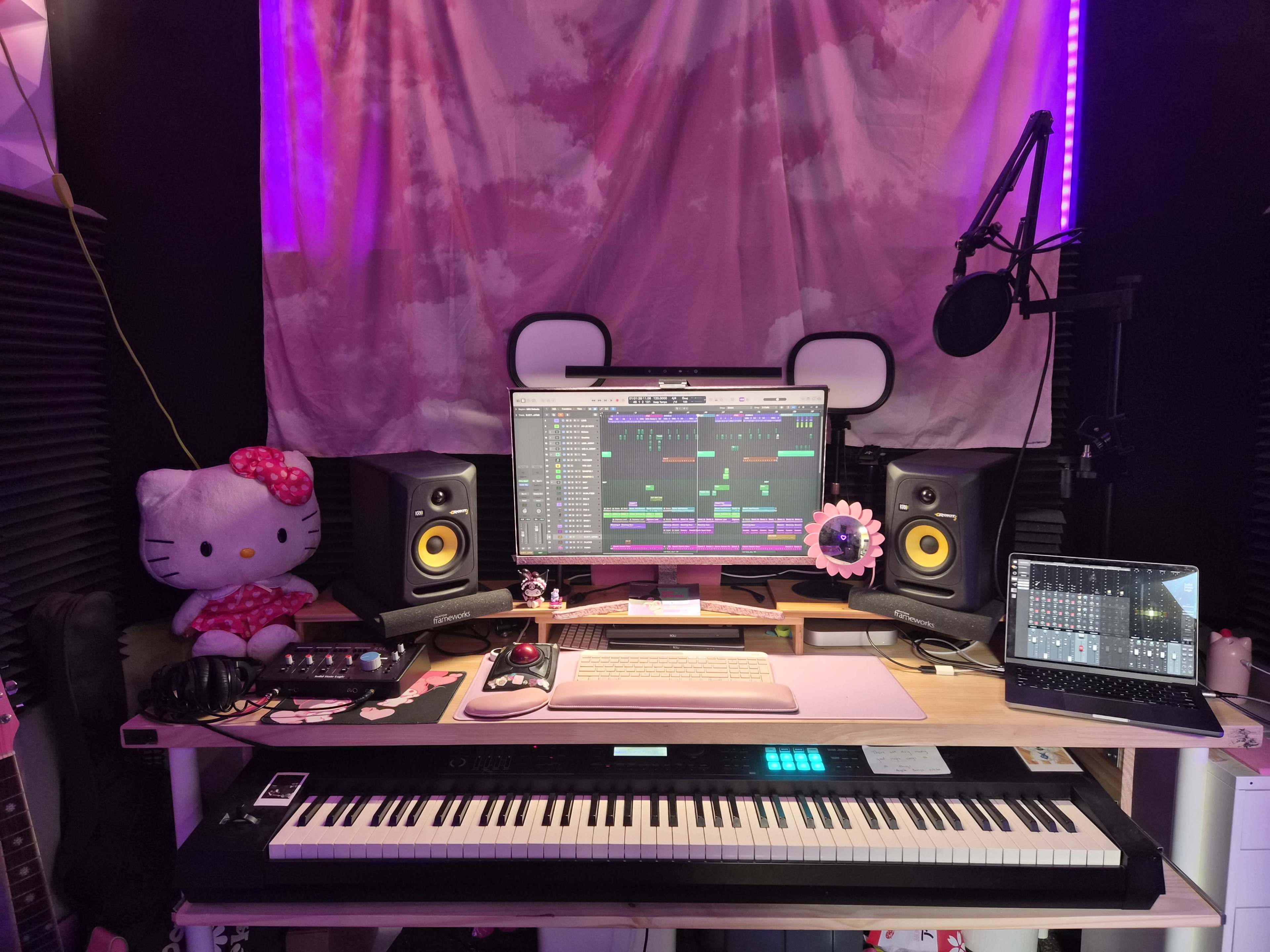 A music production setup featuring a keyboard, computer monitor displaying music software, speakers, a microphone, headphones, and decorative elements including a Hello Kitty plush toy and a sunflower.